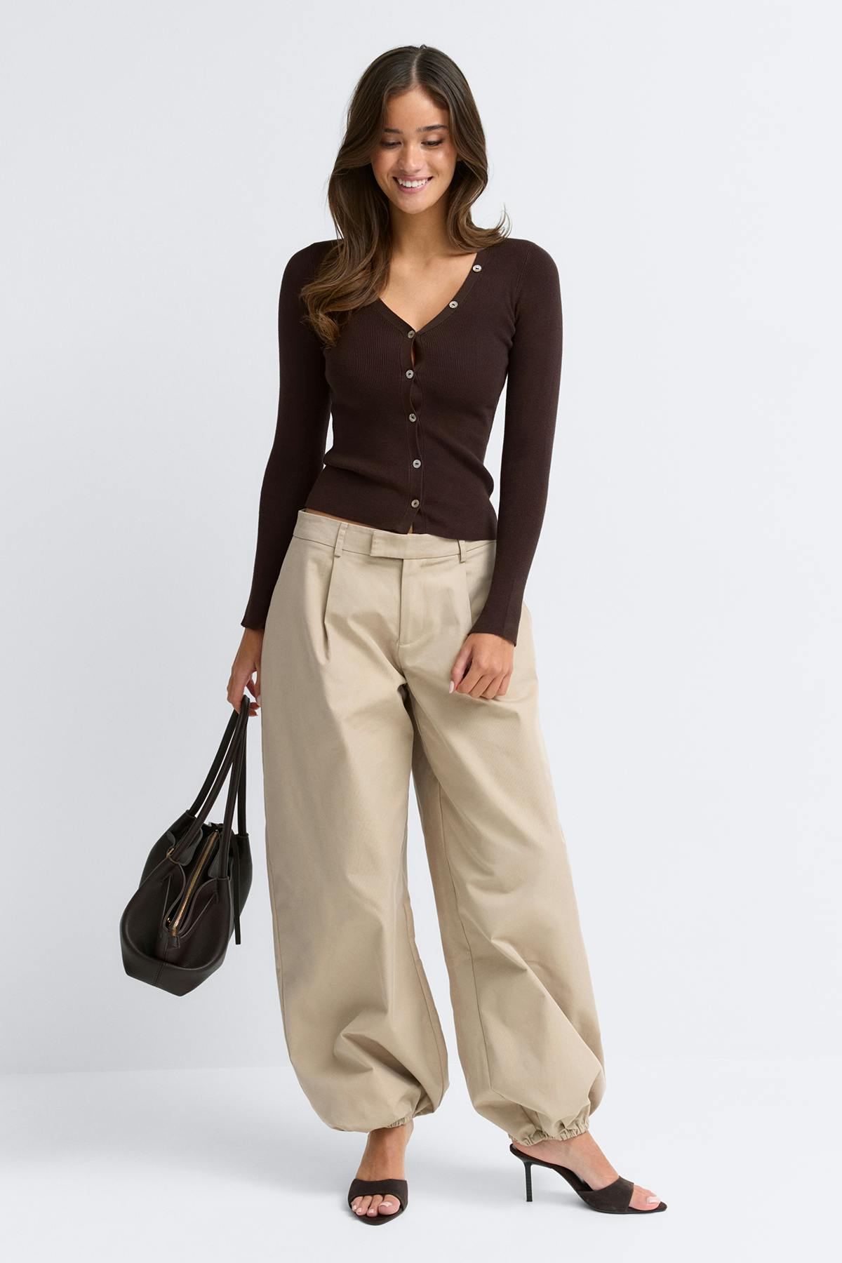 Tummanruskea uurrettu V-pääntie-neuletakki, jossa on nappikiinnitys; korkeavyötäröiset beigenväriset leveälahkeiset laskostetut housut, joissa on joustavat nilkat; tummanruskeat avokärkiset korkokenkäkengät; tummanruskea nahkakantolaukku.