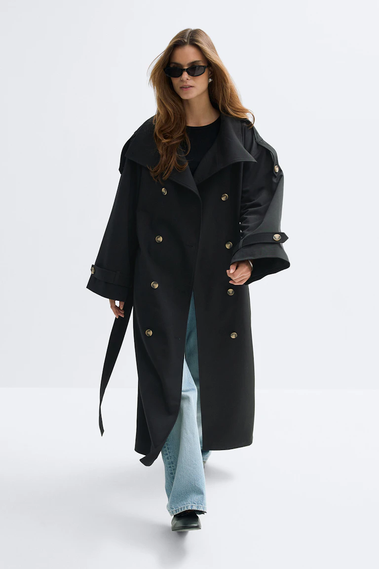 Long trench-coat noir à double boutonnage avec boutons dorés, ceinture et pattes de manches, porté sur un top noir à col ras du cou, un jean évasé délavé, des chaussures noires à bout pointu et des lunettes de soleil noires œil de chat.