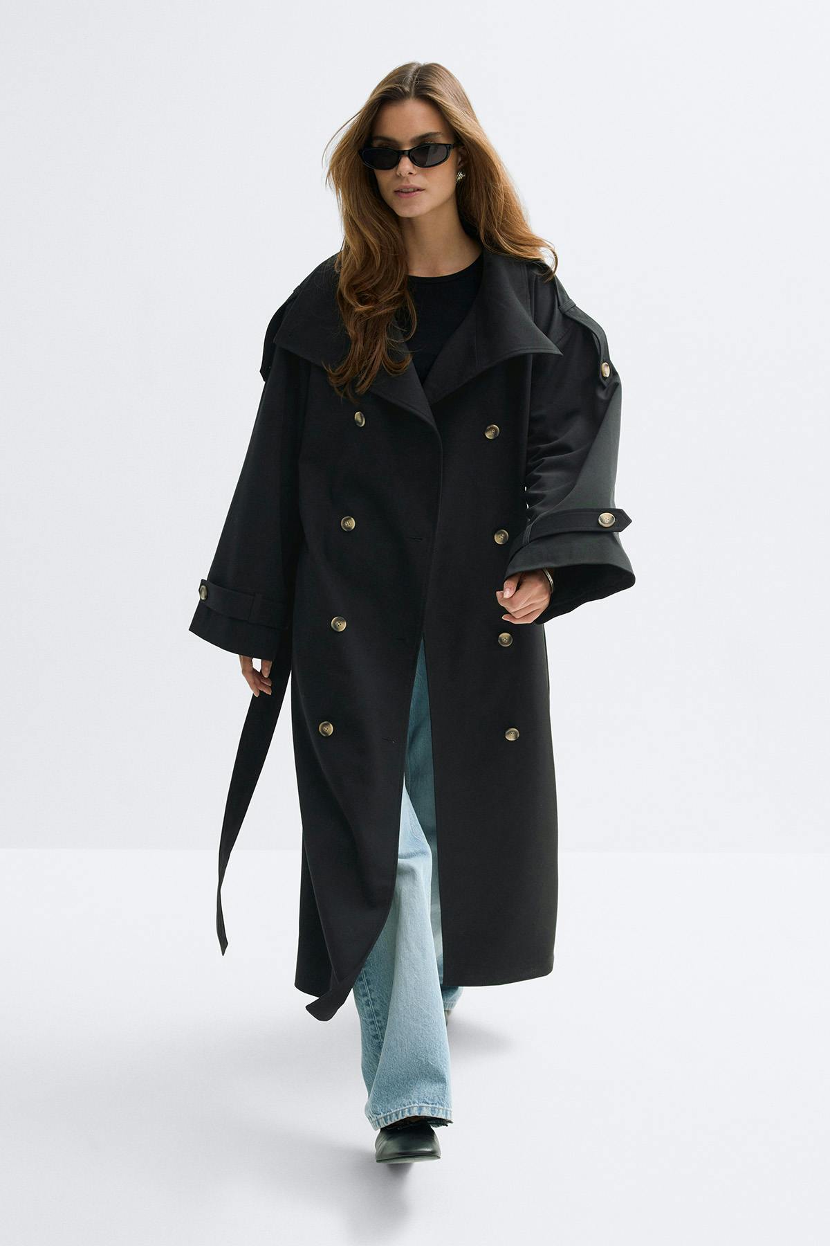 Long trench-coat noir à double boutonnage avec boutons dorés, ceinture et pattes de manches, porté sur un top noir à col ras du cou, un jean évasé délavé, des chaussures noires à bout pointu et des lunettes de soleil noires œil de chat.