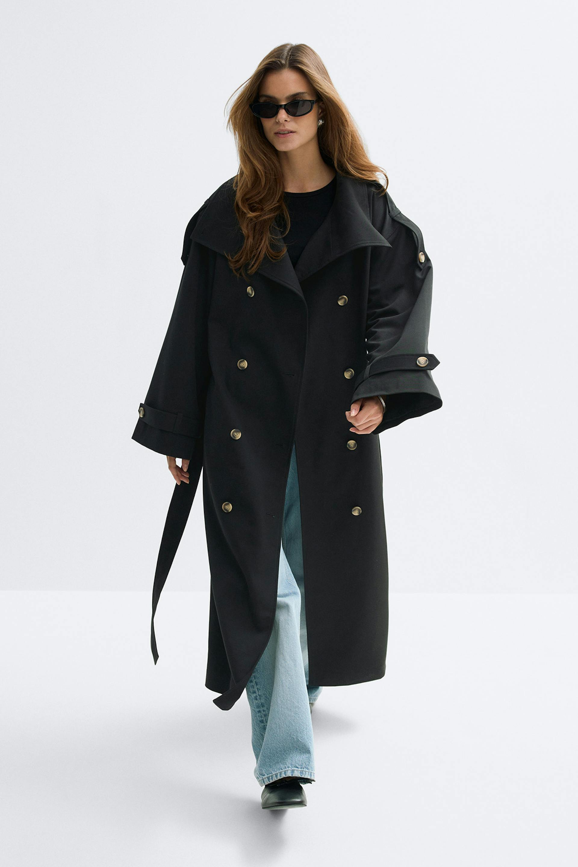 Long trench-coat noir à double boutonnage avec boutons dorés, ceinture et pattes de manches, porté sur un top noir à col ras du cou, un jean évasé délavé, des chaussures noires à bout pointu et des lunettes de soleil noires œil de chat.