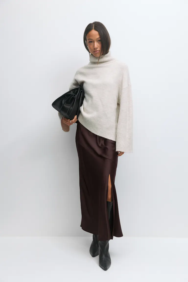 Cremefarbener Rollkragenpullover mit gerippten Bündchen und Saum, dunkelbrauner Midirock aus Satin mit Seitenschlitz, schwarze, kniehohe, spitze Lederstiefel und eine schwarze Lederhandtasche.