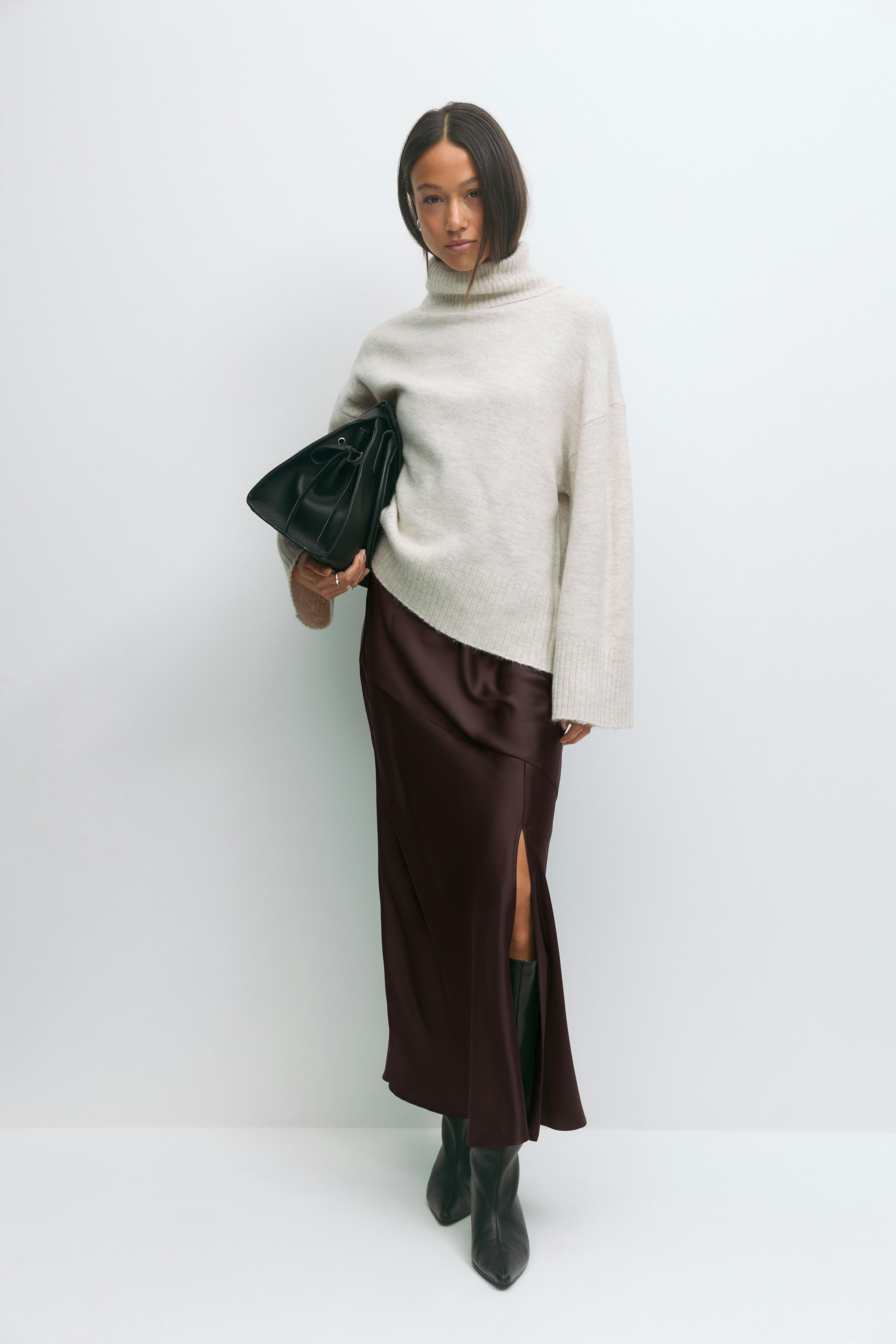 Cremefarbener Rollkragenpullover mit gerippten Bündchen und Saum, dunkelbrauner Midirock aus Satin mit Seitenschlitz, schwarze, kniehohe, spitze Lederstiefel und eine schwarze Lederhandtasche.