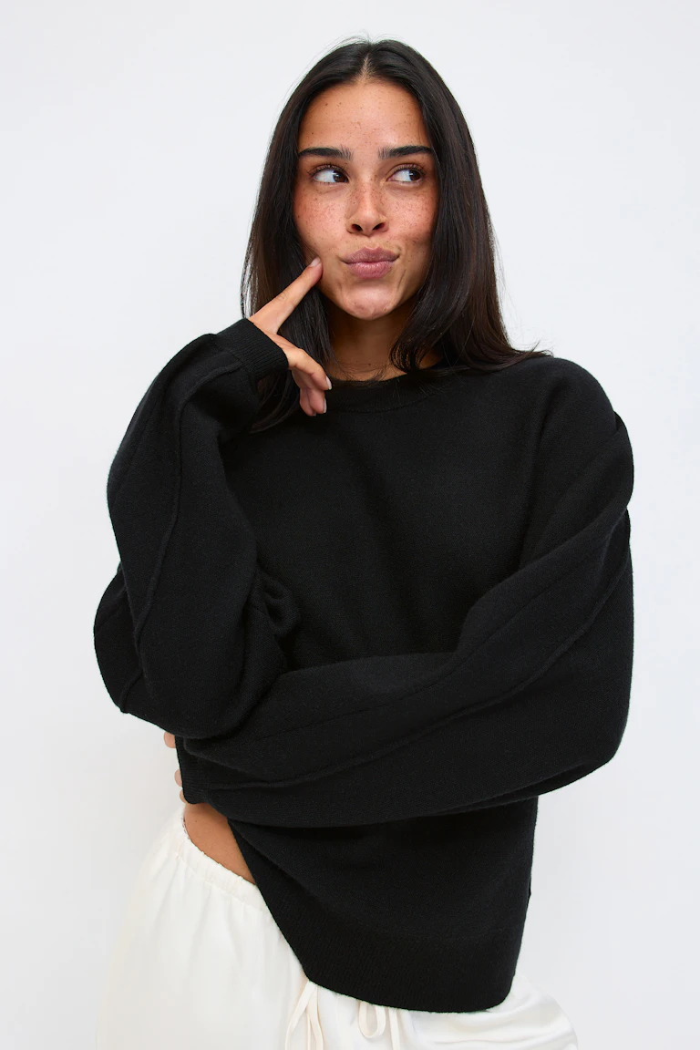 Schwarzer Oversize-Strickpullover mit Rundhalsausschnitt und gerippten Bündchen und Saum, kombiniert mit einer weißen Hose mit Kordelzug.