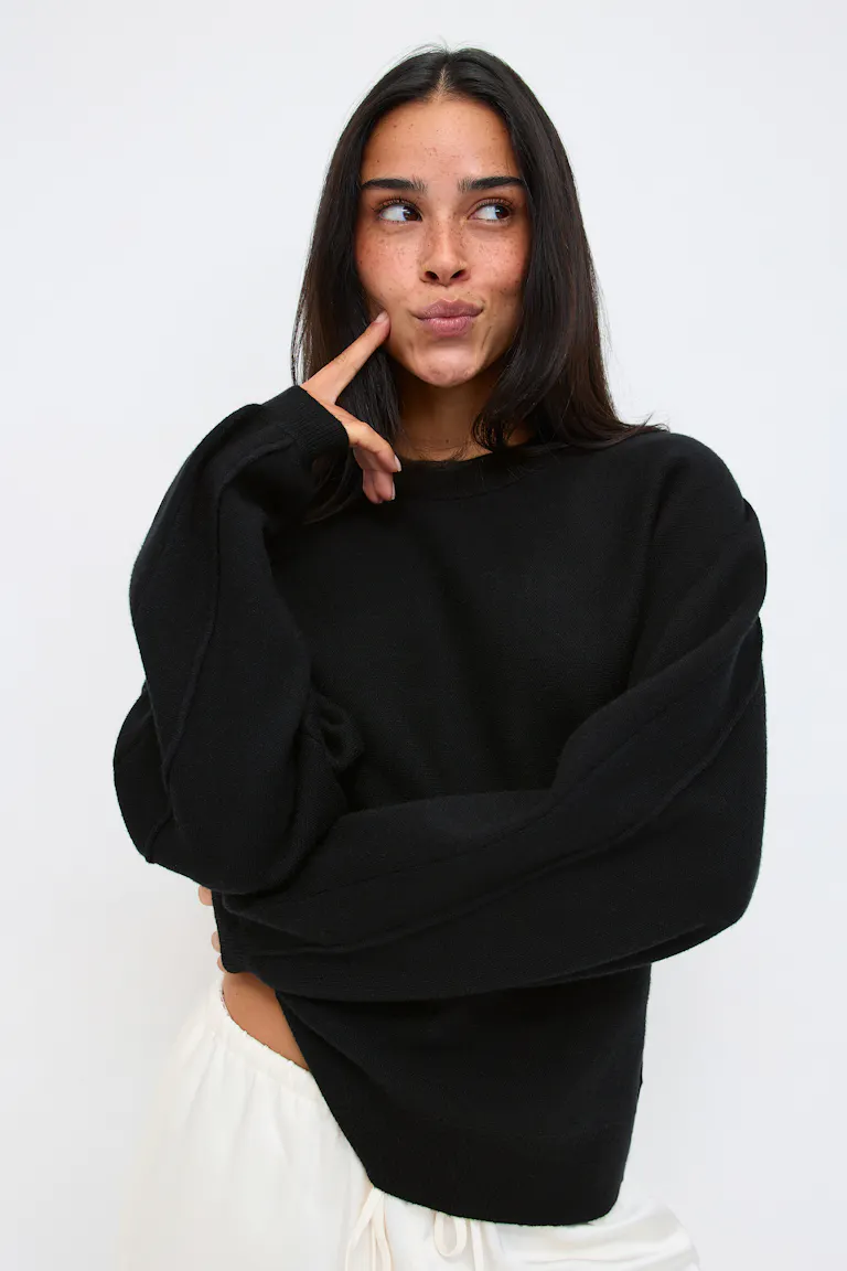 Schwarzer Oversize-Strickpullover mit Rundhalsausschnitt und gerippten Bündchen und Saum, kombiniert mit einer weißen Hose mit Kordelzug.