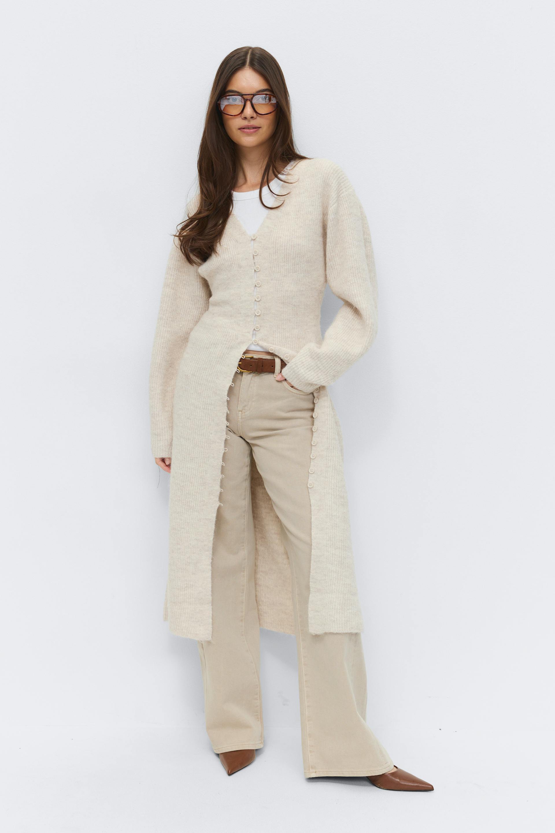 Beige long ribbed button-front duster cardigan over a white crew-neck top, paired with beige wide-leg trousers, a brown leather belt, brown pointed-toe boots, and tortoiseshell glasses.