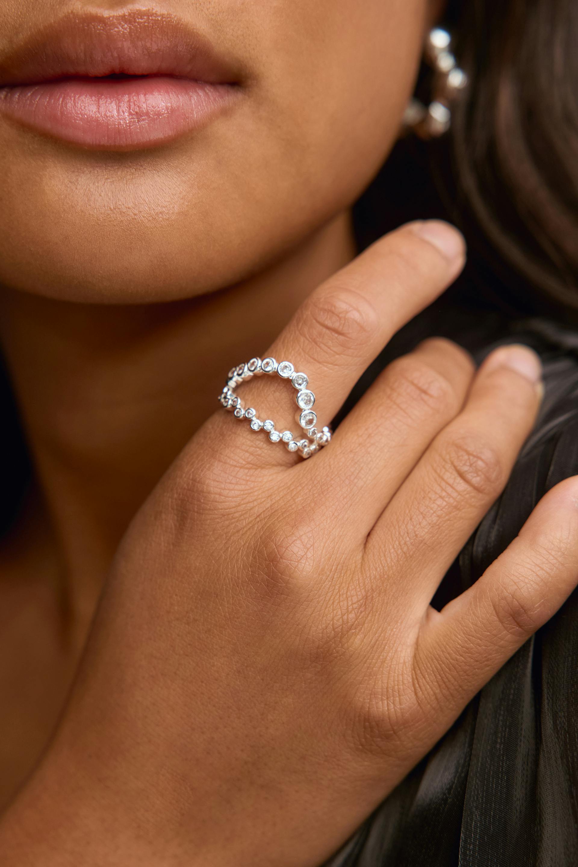 Silver double-band ring with graduated round clear stones worn on the index finger; polished silver hoop earring visible.