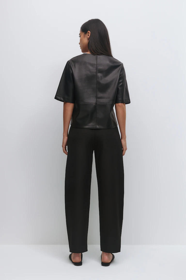 Schwarzes kurzärmeliges, kastenförmiges Oberteil aus Leder mit Nahtdetails, kombiniert mit einer schwarzen Hose mit hoher Taille und schwarzen Slide-Sandalen.