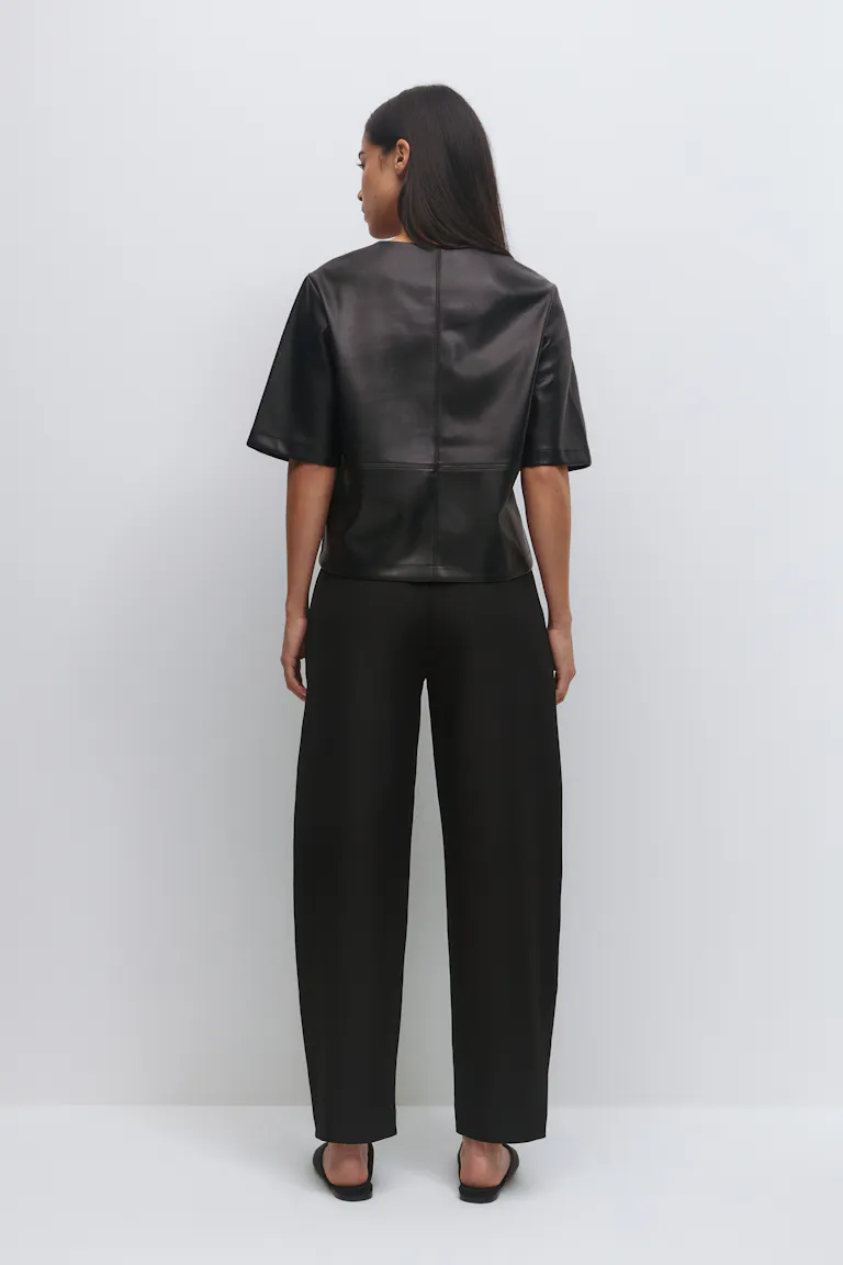 Schwarzes kurzärmeliges, kastenförmiges Oberteil aus Leder mit Nahtdetails, kombiniert mit einer schwarzen Hose mit hoher Taille und schwarzen Slide-Sandalen.