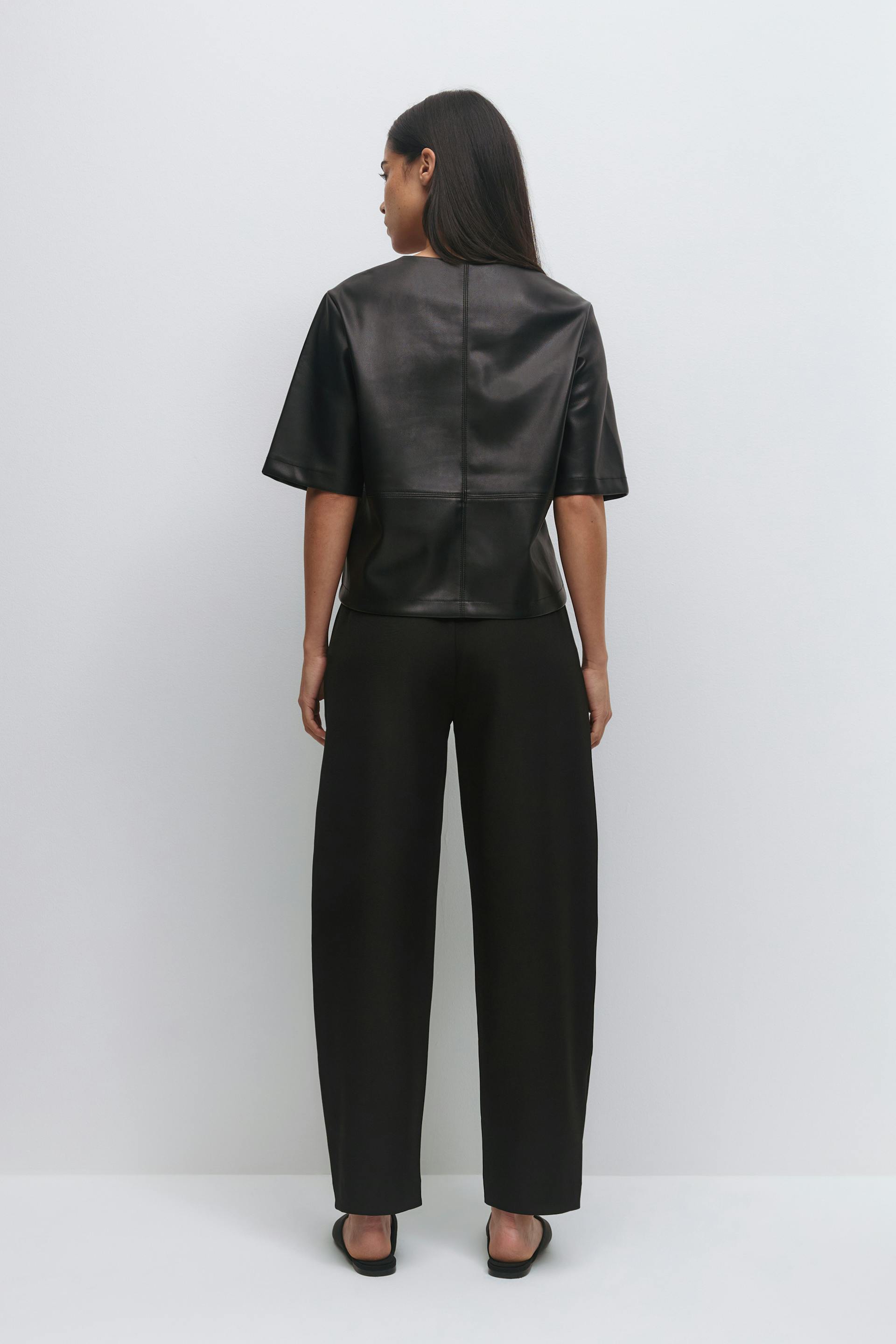 Black short-sleeve leather boxy top with seam detailing, paired with high‑waisted wide‑leg black trousers and black slide sandals.