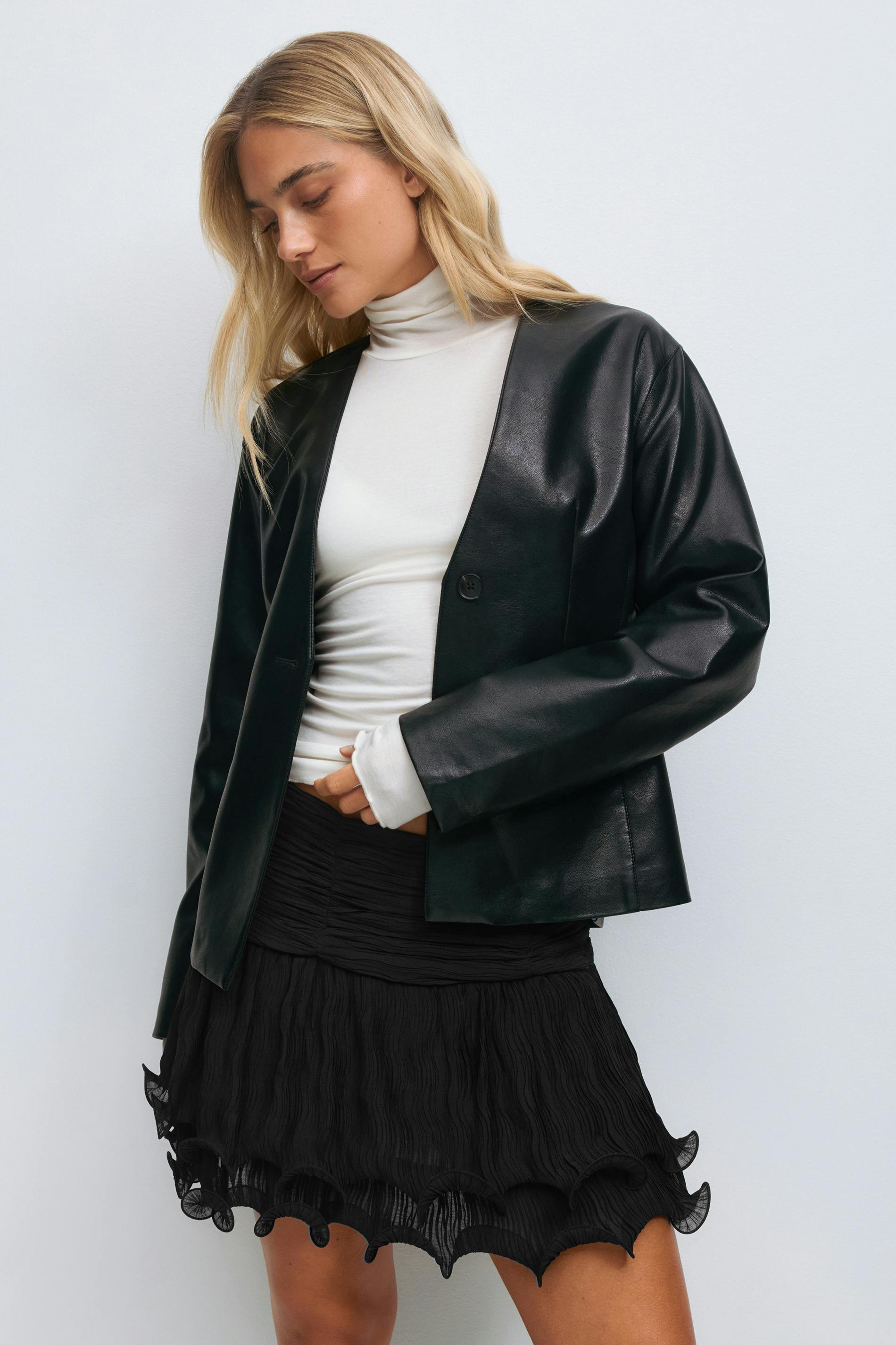 Black pleated long-sleeve blouse with tie-neck and ruffled cropped hem, paired with a matching high-waist pleated mini skirt with scalloped ruffle trim, sheer black tights, black strappy heeled sandals with gold circular hardware, and gold hoop earrings.
