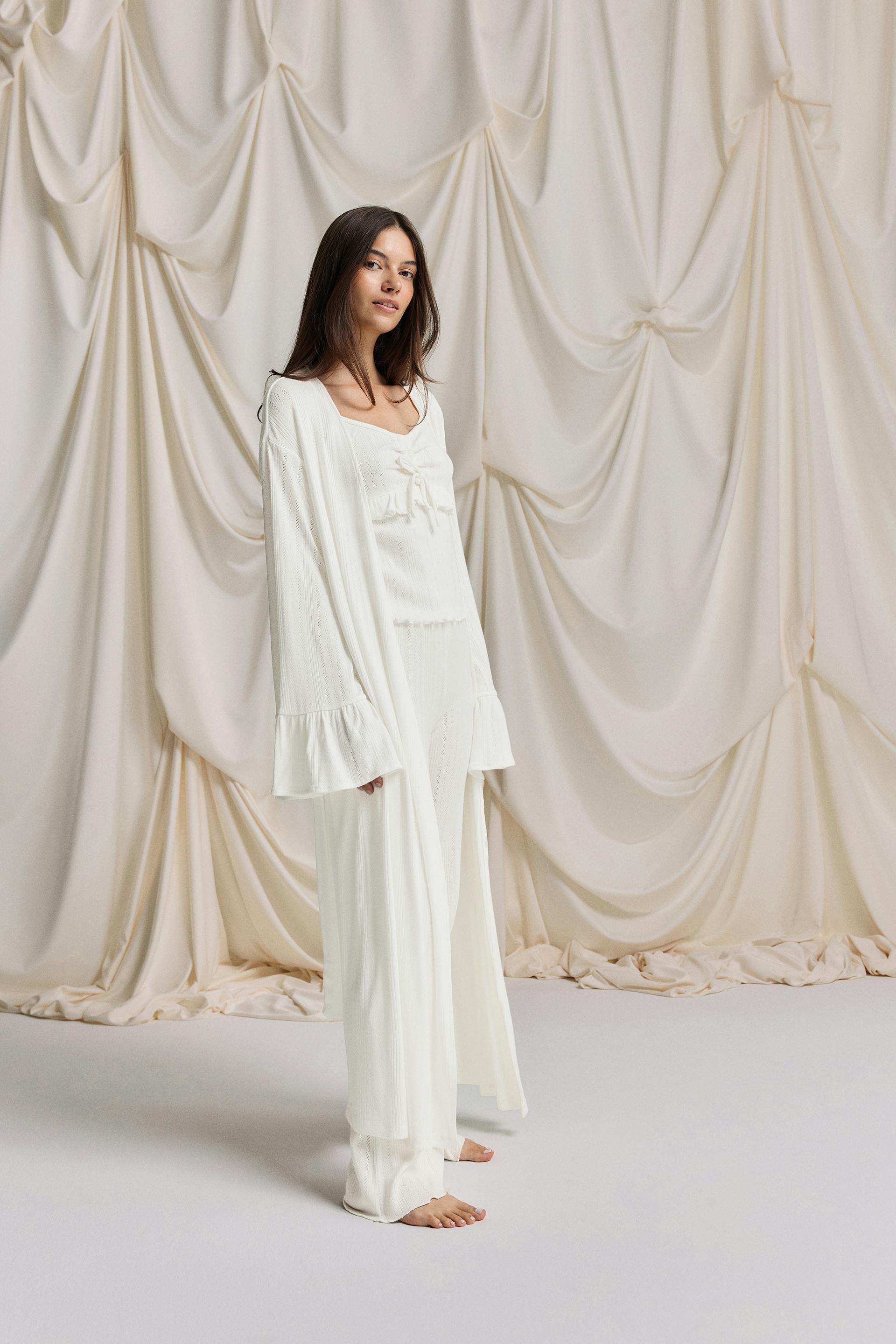 Long white longline knit robe with waist tie and flared bell sleeves, worn over a lace‑up white camisole and matching wide‑leg knit trousers.