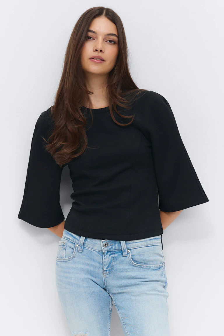 Zwarte getailleerde top met ronde hals en ellebooglange uitlopende mouwen, gecombineerd met een slanke jeans in lichte wassing met subtiele distressing.