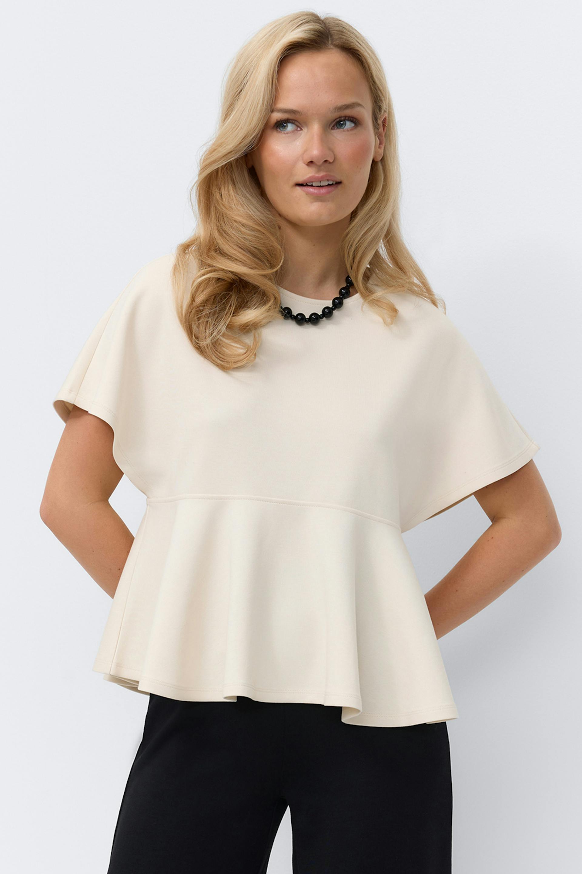 Top peplum à manches courtes de couleur crème et à encolure ronde, associé à un pantalon droit noir et à un collier de perles noires.