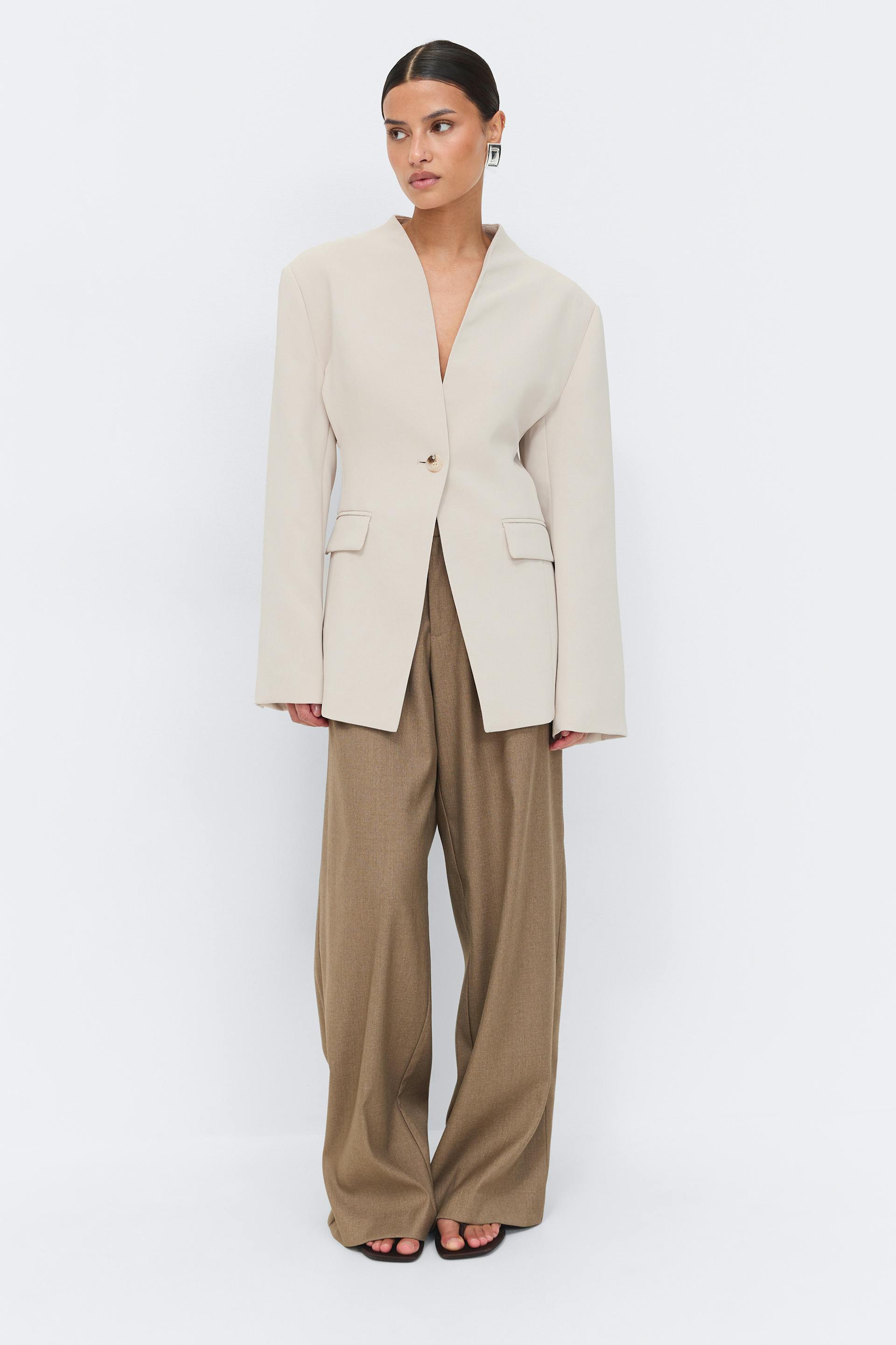 Light beige single-button tailored blazer with long sleeves and flap pockets, paired with wide-leg brown trousers and black open-toe sandals.