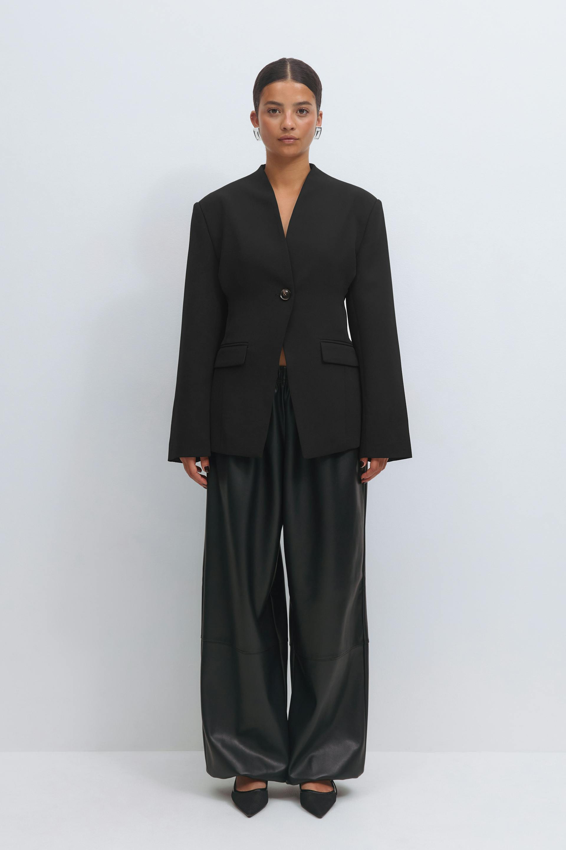 Black single-button tailored blazer with a deep V neckline and flap pockets, paired with high-waisted wide-leg black faux-leather trousers and pointed-toe black pumps.
