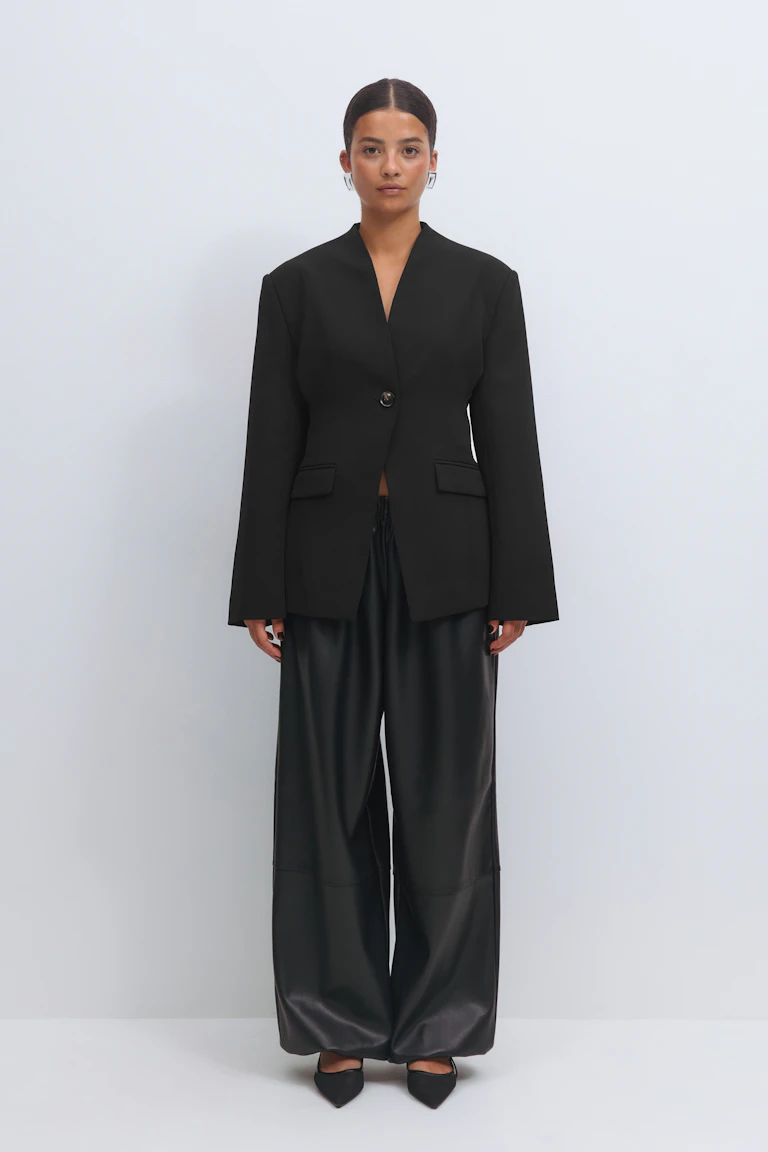 Schwarzer, taillierter Blazer mit tiefem V-Ausschnitt und Klappentaschen, kombiniert mit einer schwarzen Kunstlederhose mit weitem Bein und spitzen schwarzen Pumps.