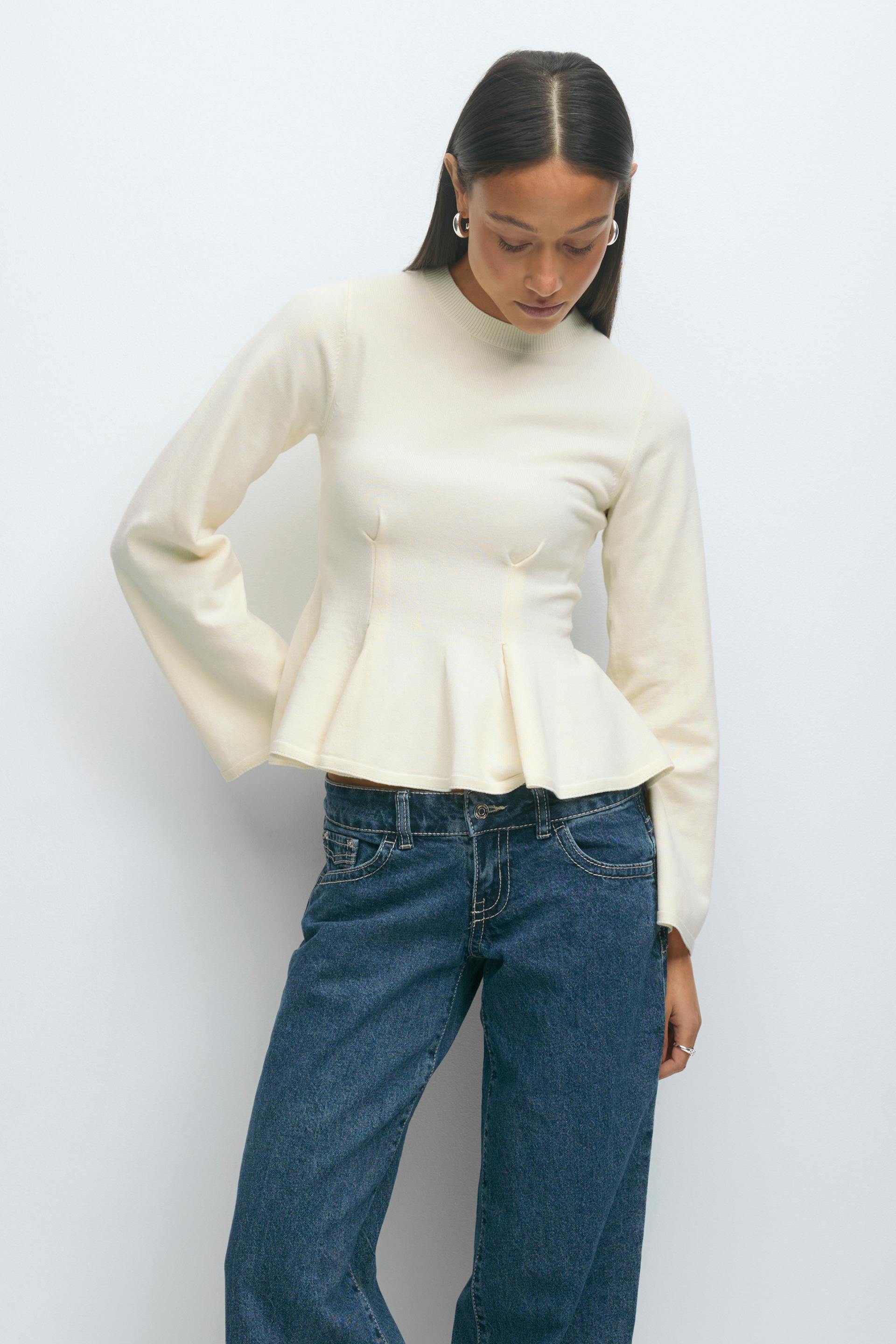 Cream long-sleeve peplum knit top with high neckline and subtle bust seaming, paired with dark blue straight-leg jeans with contrast stitching and silver hoop earrings.