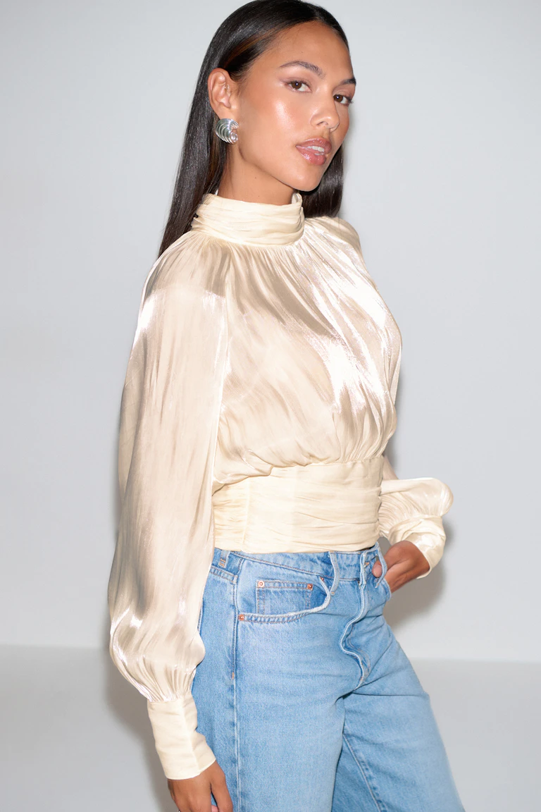 Langärmelige Bluse aus cremefarbenem Satin mit hohem Halsausschnitt, geraffter Taille und taillierten Manschetten, kombiniert mit hellen High-Waist-Jeans und silbernen Statement-Ohrringen.