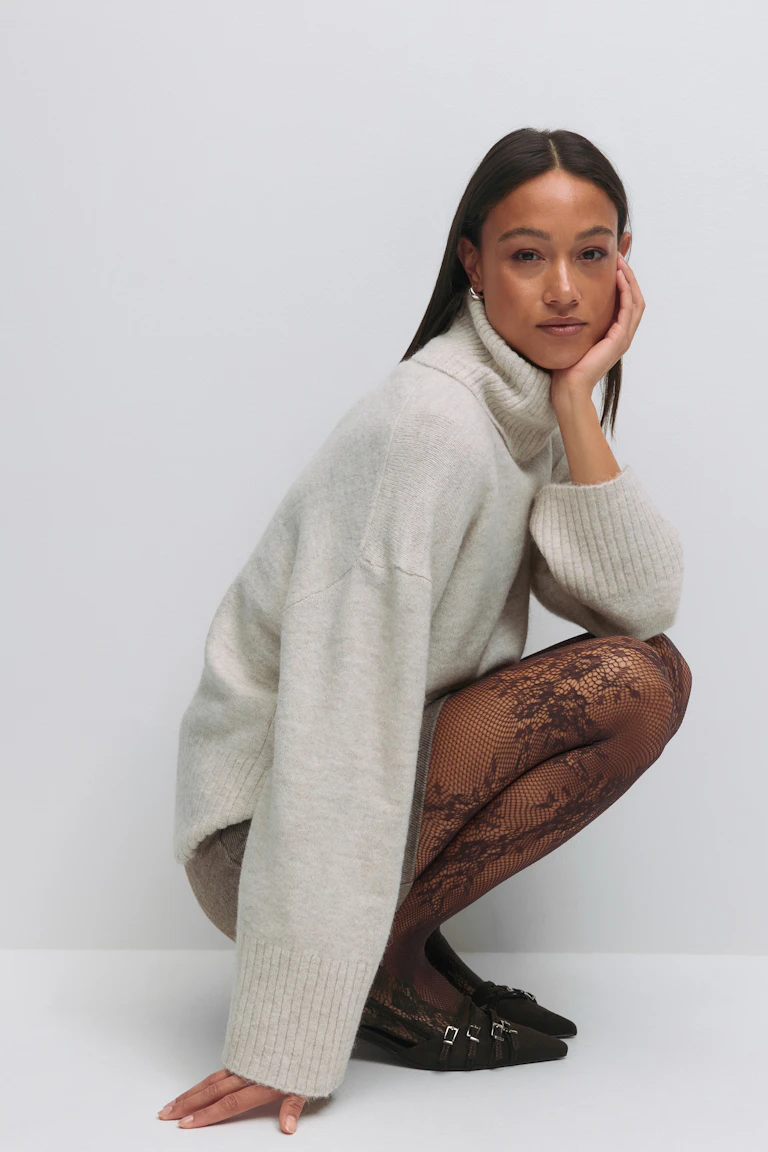 Cremefarbener, übergroßer, gerippter Rollkragenpullover, schwarze Netzstrumpfhose mit Blumenmuster und schwarze, spitze Schuhe mit mehreren Schnallenriemen.