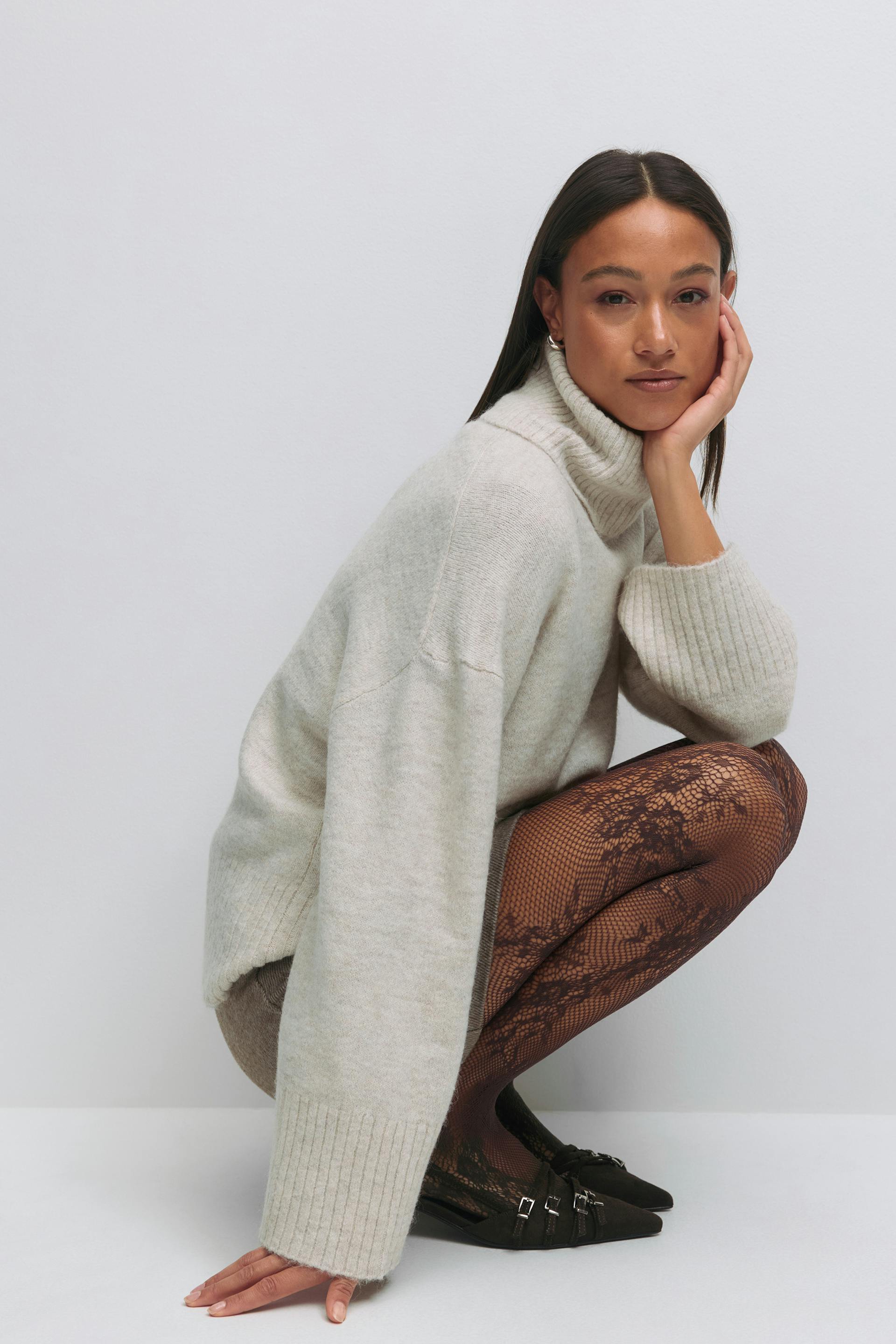 Cremefarbener, übergroßer, gerippter Rollkragenpullover, schwarze Netzstrumpfhose mit Blumenmuster und schwarze, spitze Schuhe mit mehreren Schnallenriemen.