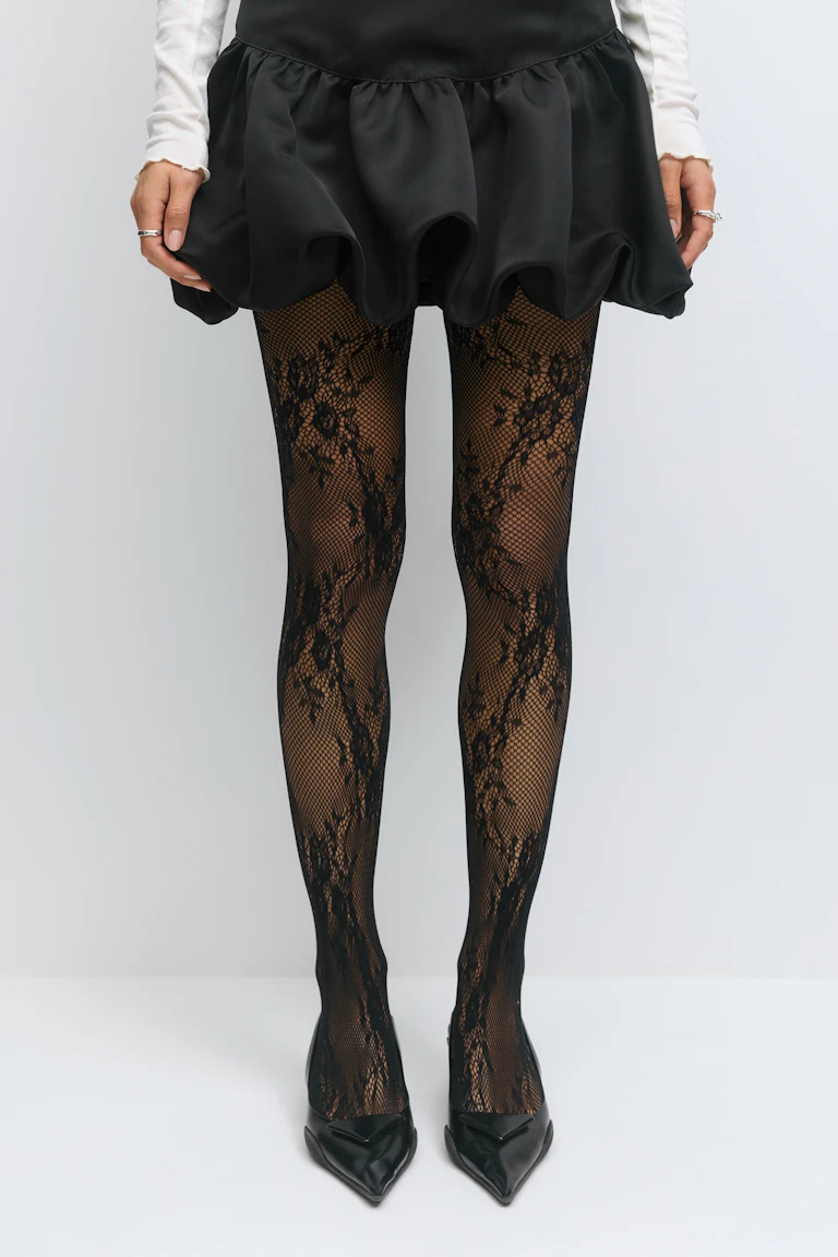 Model wearing a black ruffled mini skirt, white long-sleeve top (sleeves visible), black lace-pattern fishnet tights, and black pointed-toe heels.