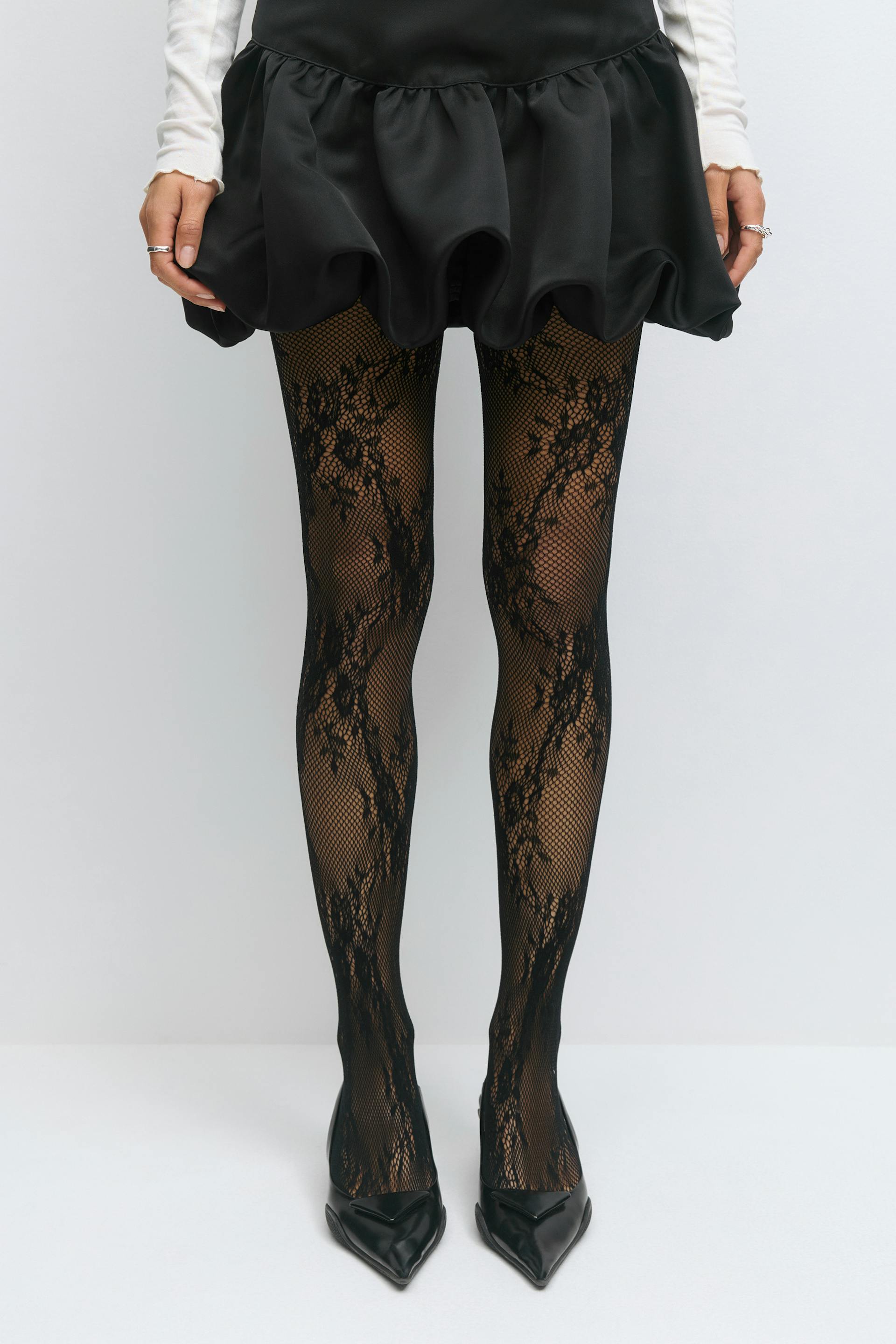 Das Modell trägt einen schwarzen gerüschten Minirock, ein weißes langärmeliges Oberteil (Ärmel sichtbar), eine schwarze Netzstrumpfhose mit Spitzenmuster und schwarze spitze Absätze.