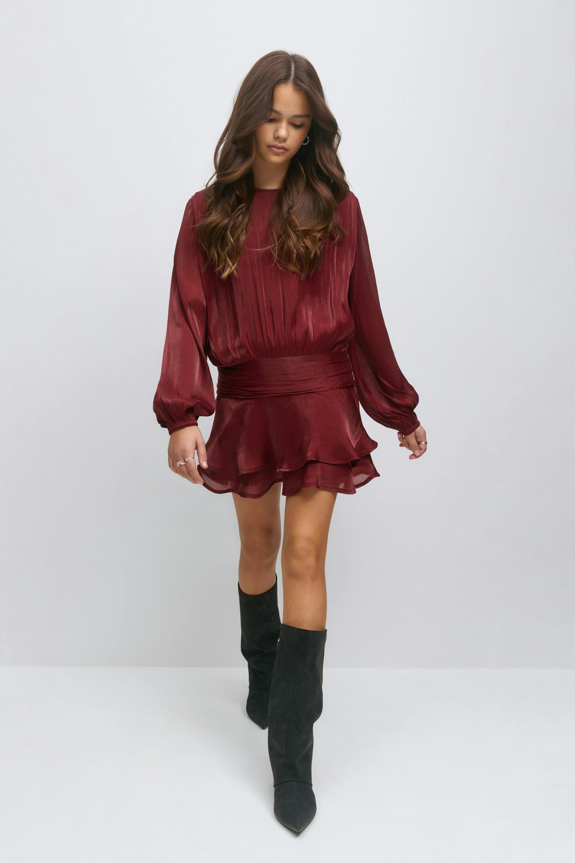 Model dressed in a burgundy long-sleeve satin mini dress with a pleated bodice, wide cinched waistband and layered ruffle hem, paired with black knee-high pointed-toe suede boots.
