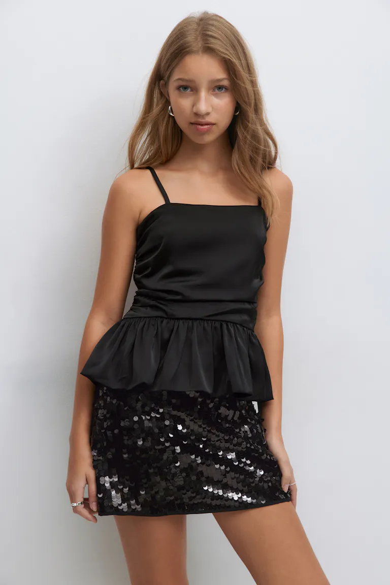 Sort satin camisole med tynde stropper, firkantet halsudskæring og peplum flæse, parret med en sort paillet mini nederdel og små sølv hoop øreringe.