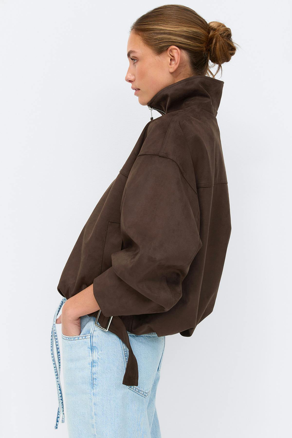 Veste zippée surdimensionnée en suédine marron foncé avec col montant, fermeture à glissière argentée sur le devant, poches latérales et pattes à boucles réglables à l'ourlet, portée avec un pantalon noir.
