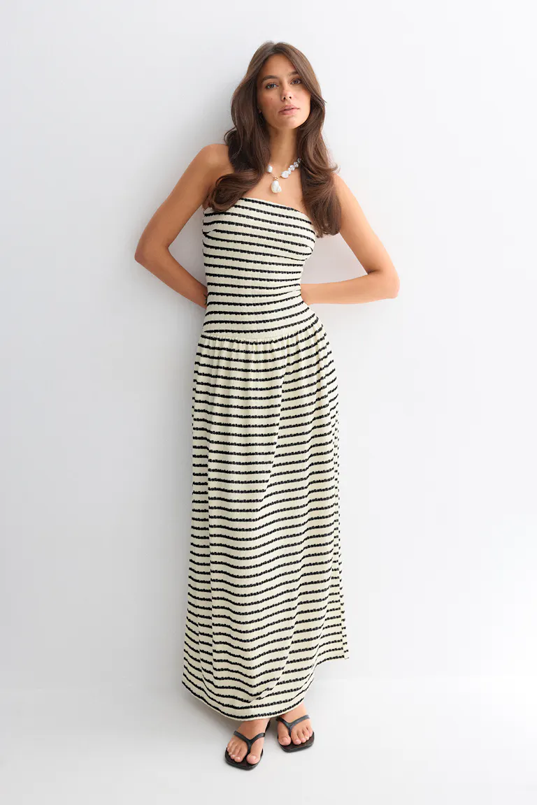 Trägerloses creme-schwarzes, horizontal gestreiftes Maxikleid mit tailliertem, gesmoktem Oberteil, kombiniert mit schwarzen Zehenstegsandalen und einer klobigen Perlenkette.