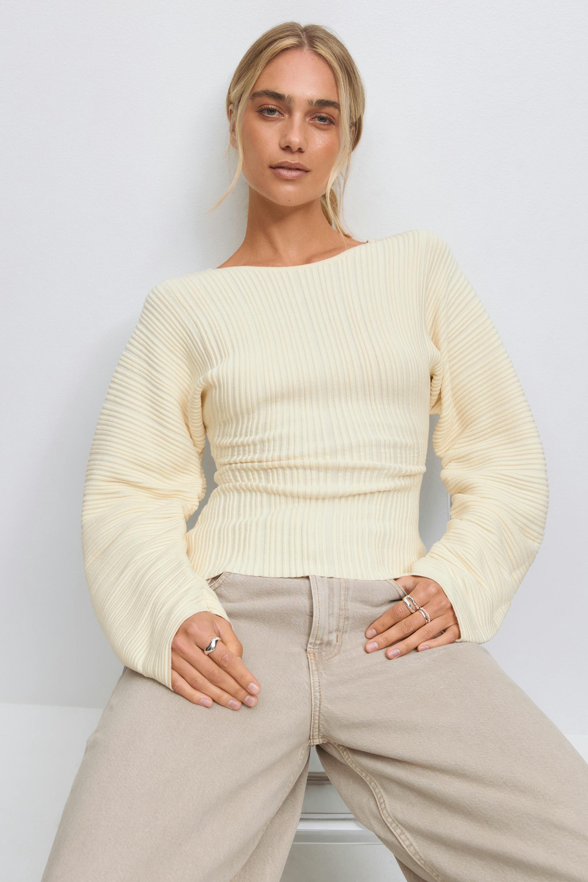 Model wearing a cream ribbed boat-neck sweater with a fitted waist and voluminous long sleeves, paired with high‑waisted beige wide‑leg jeans.