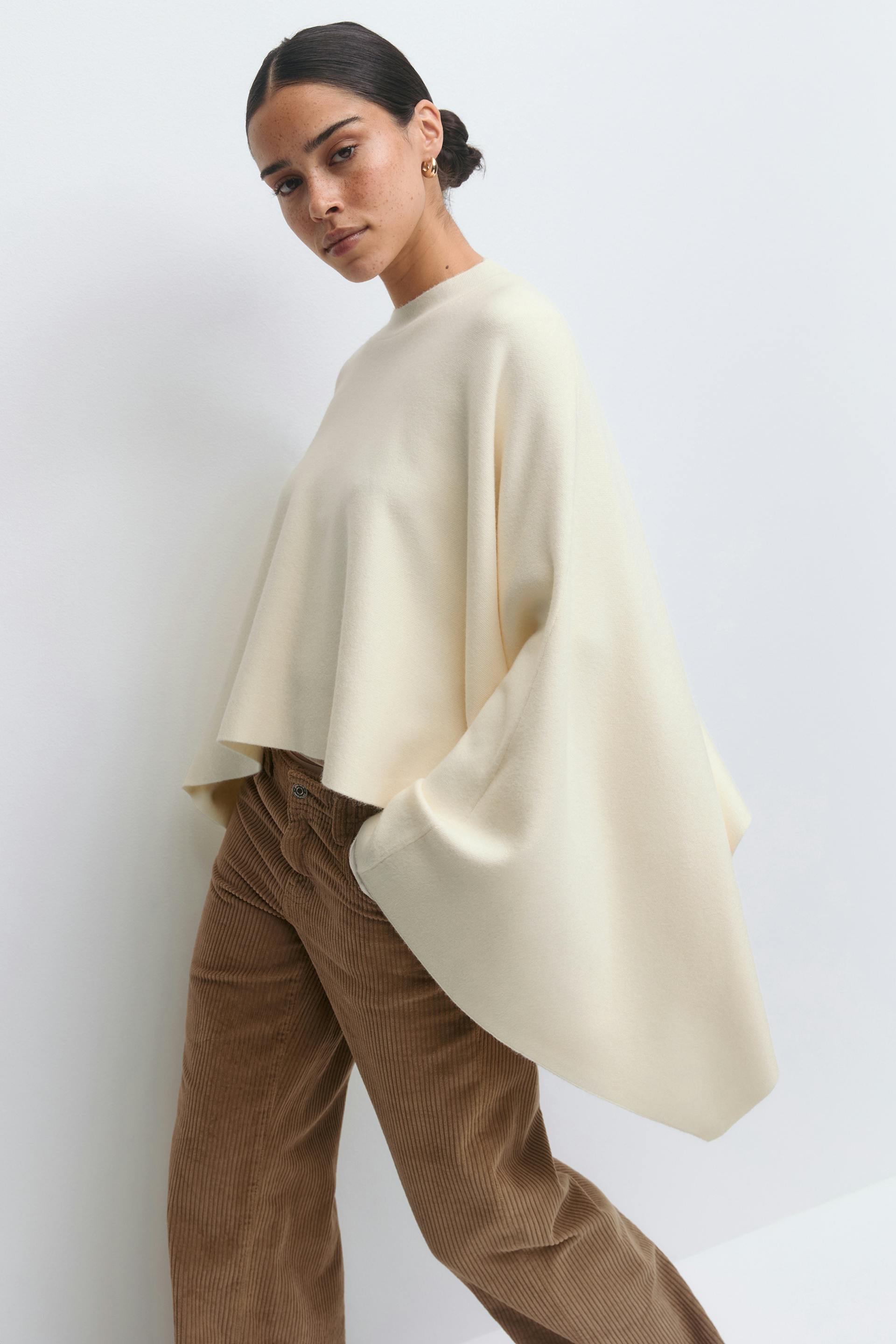 Overdimensjonert genser i kremfarget cape-stil med høy rund hals, kombinert med en brun kordfløyelsbukse.