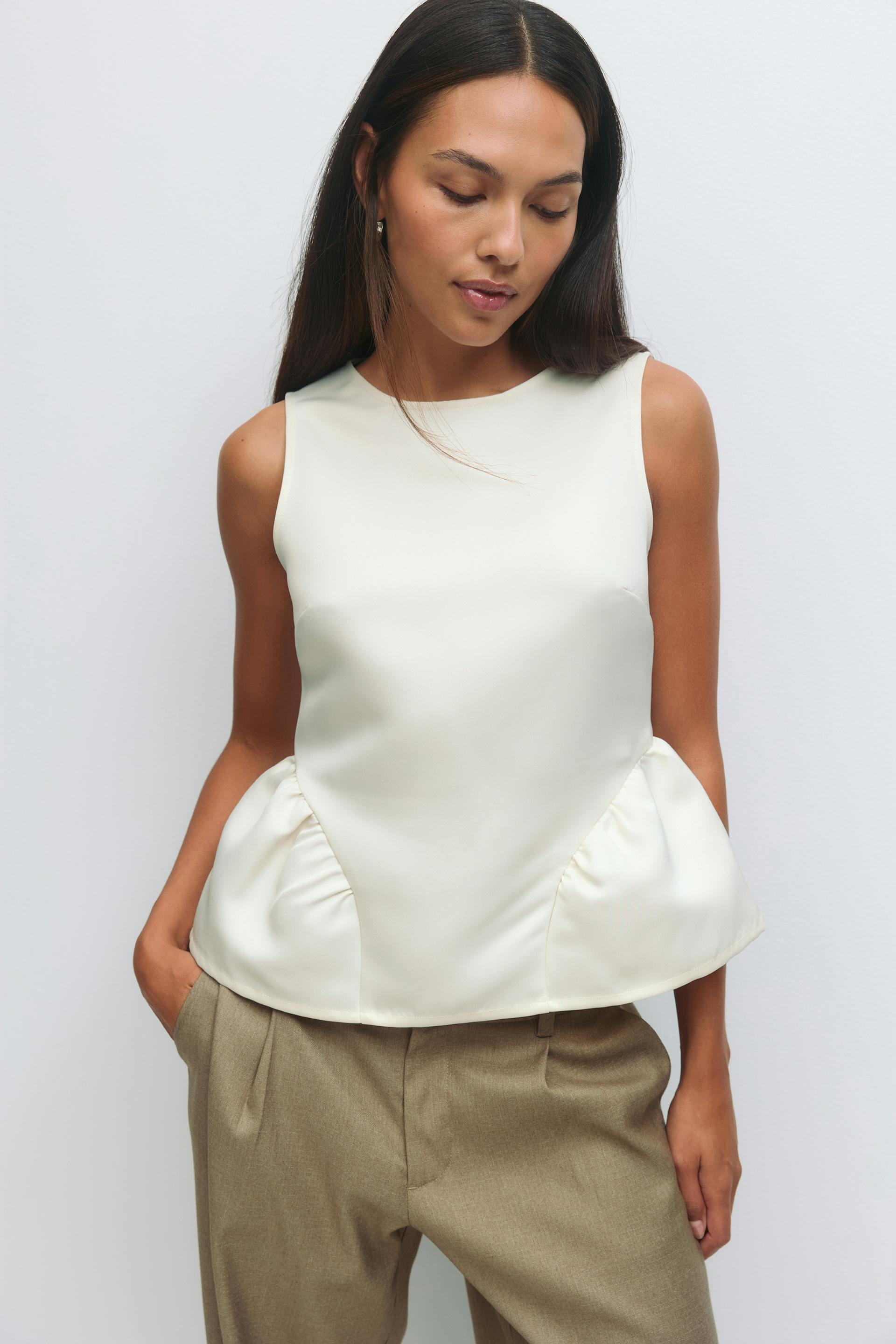 Mouwloze ivoorkleurige satijnen peplum top met een hoge ronde halslijn, gecombineerd met een beige broek met plooien.