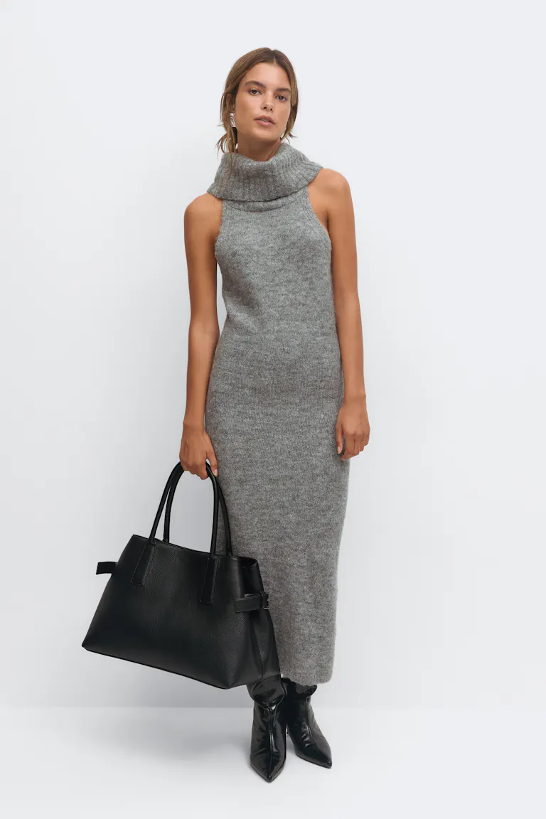 Mouwloze grijze gebreide midi-jurk met een omgeslagen col, gestyled met zwarte enkellaarsjes met spitse teen, een grote zwarte leren tas en zilveren oorbellen.