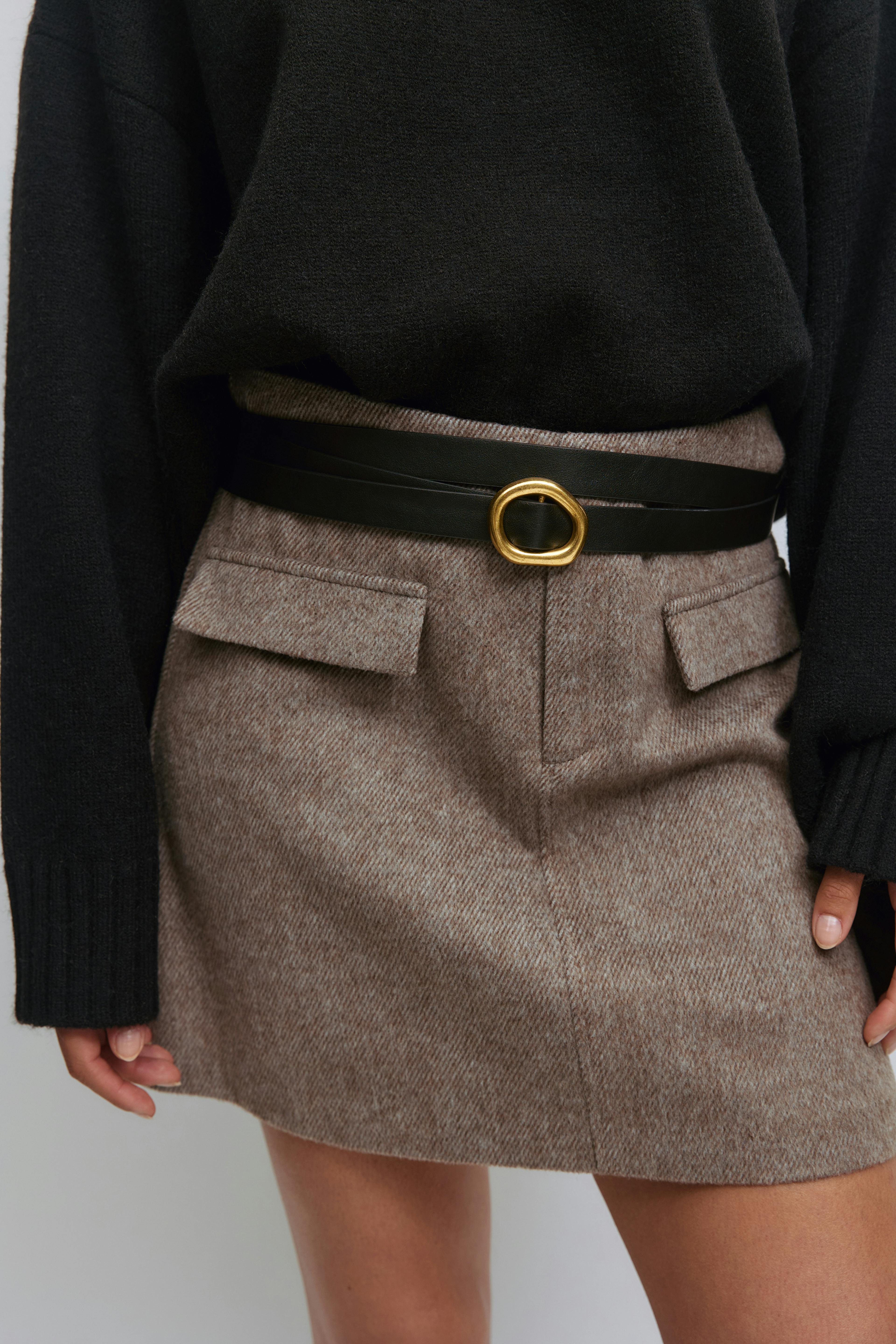 Pull noir en maille à manches longues associé à une mini-jupe en laine taupe à poches à rabats, cintrée par une ceinture en cuir noir à double enroulement et boucle ovale dorée.