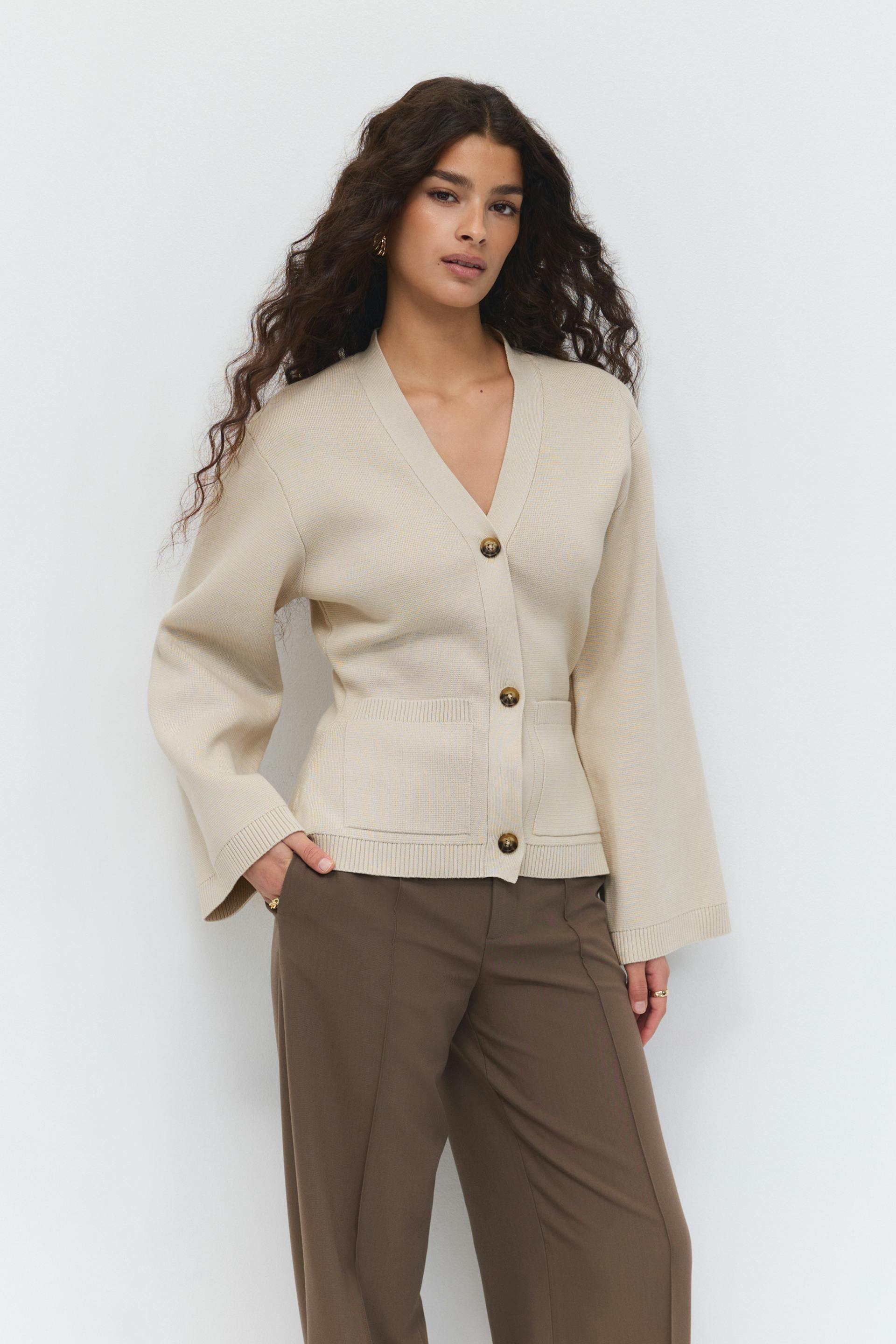Cardigan en maille beige à col en V, larges manches longues, boutons en écaille et poches plaquées sur le devant, associé à un pantalon large marron à taille haute.