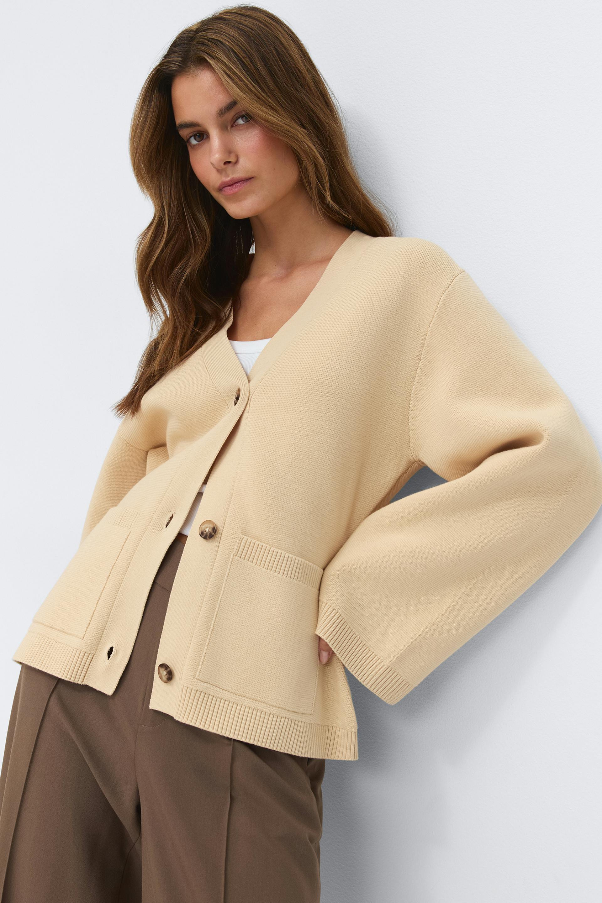 Beige oversized V‑neck button‑front cardigan with patch pockets worn over a white top, paired with brown tailored trousers.