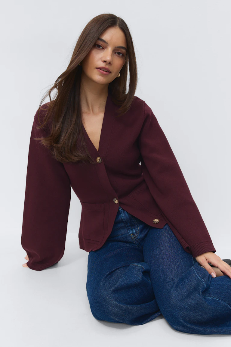 Burgunderfarbene Strickjacke mit V-Ausschnitt und Knopfleiste, langen Ärmeln und aufgesetzten Taschen auf der Vorderseite, kombiniert mit dunkelblauen, weit geschnittenen Jeans.