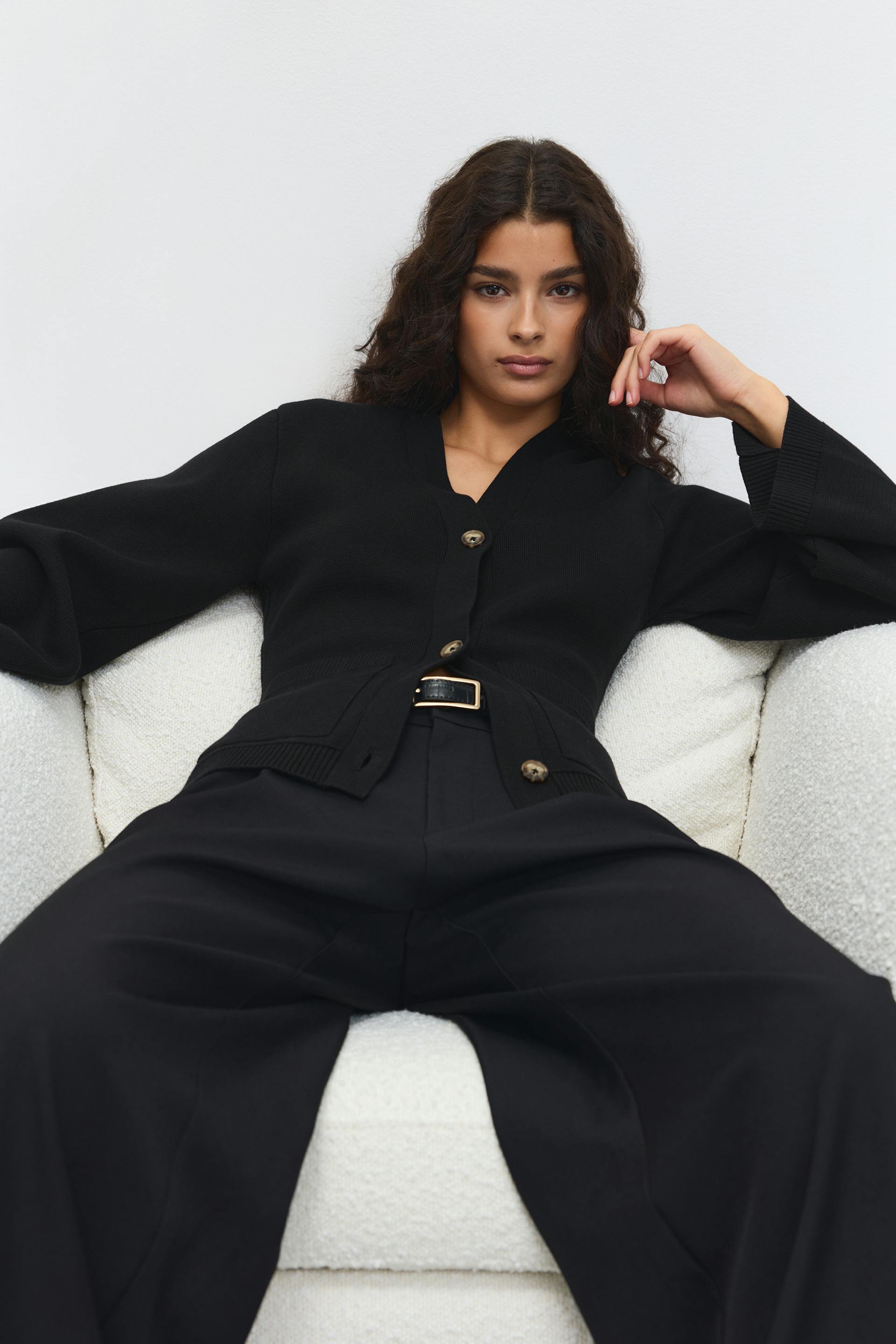 Cardigan noir boutonné à col en V avec poignets et ourlets côtelés, associé à un pantalon noir à taille haute et large et à une ceinture en cuir noir à boucle rectangulaire dorée.