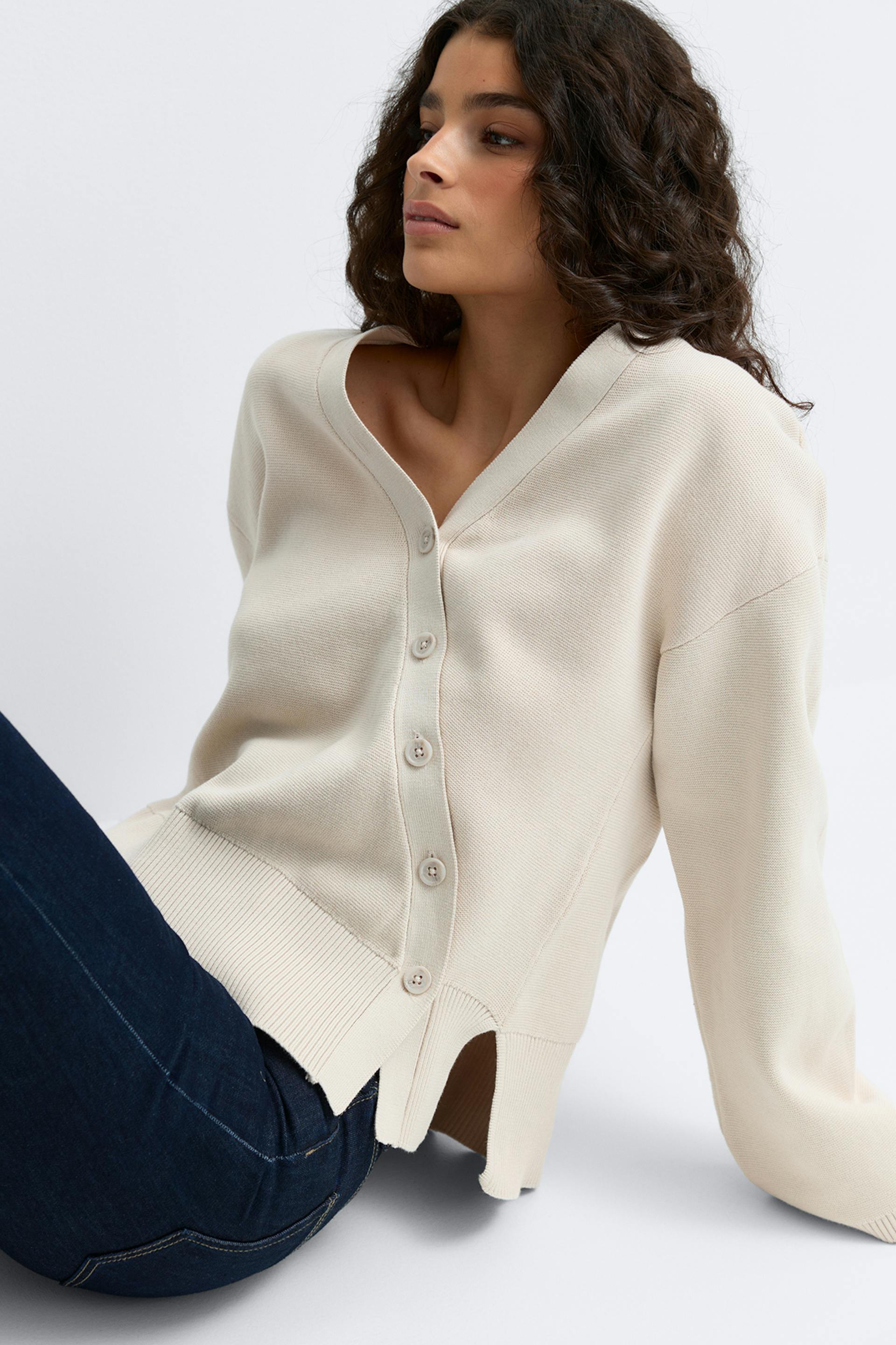 Kermanvärinen V-pääntie, napitettava hienoneulottu neuletakki, jossa on pitkät hihat, uurrettu helma ja sivuhalkiot, yhdistettynä tumman indigonvärisiin farkkuihin.