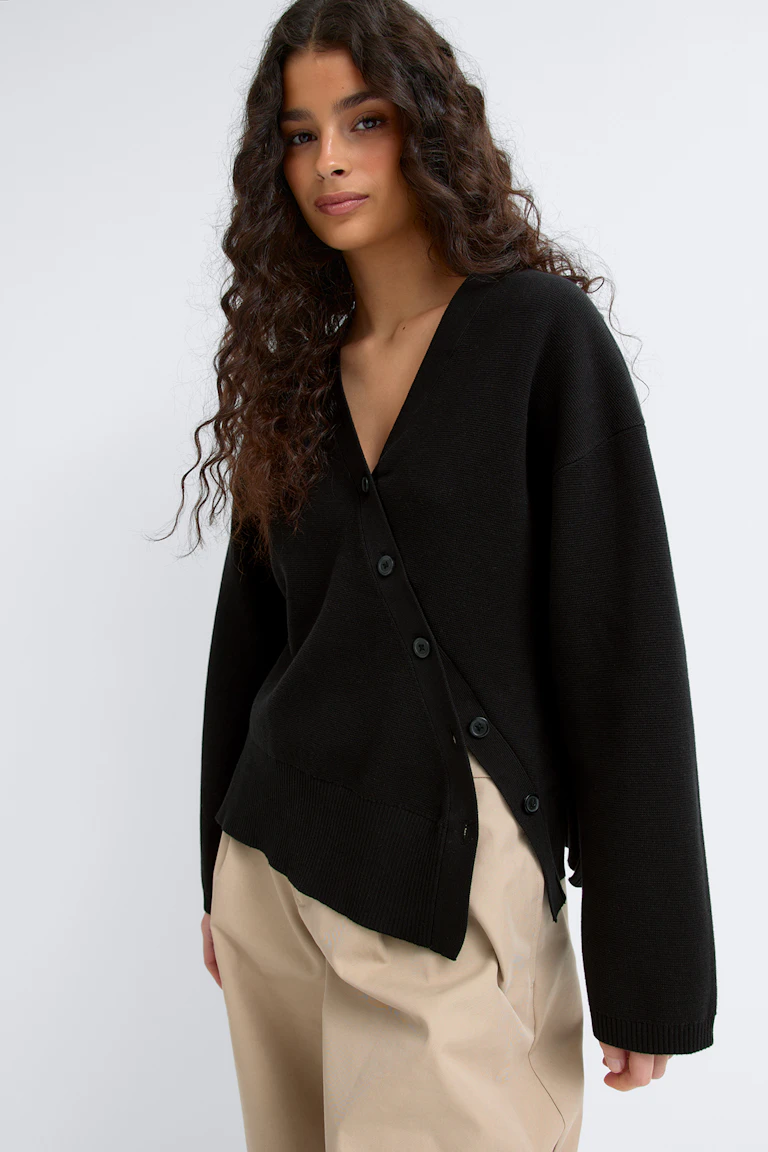Zwart vest met lange mouwen en V-hals, geknoopt met een asymmetrische knoopsluiting aan de zijkant en geribde zoom, gecombineerd met een beige broek met wijde pijpen.