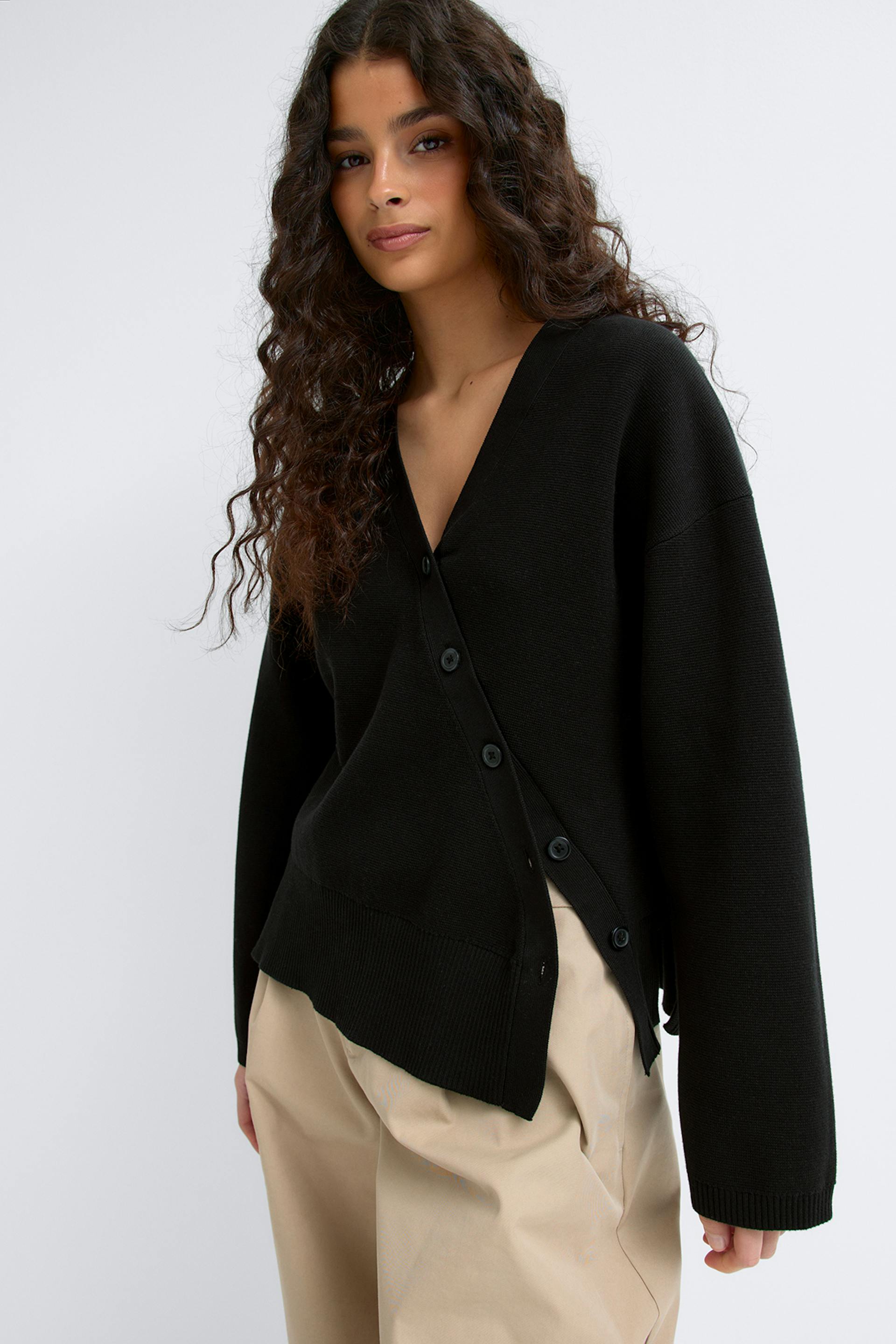 Musta V-pääntie, pitkähihainen, napitettu neuletakki, jossa on epäsymmetrinen sivunappikiinnitys ja uurrettu helma, yhdistettynä beigeen leveälahkeisiin housuihin.