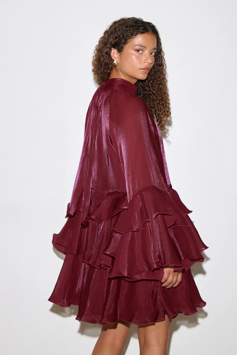 Model wearing a burgundy, satin‑finish mini dress with a high neckline, long voluminous sleeves and multiple tiered ruffle skirt, paired with gold hoop earrings.