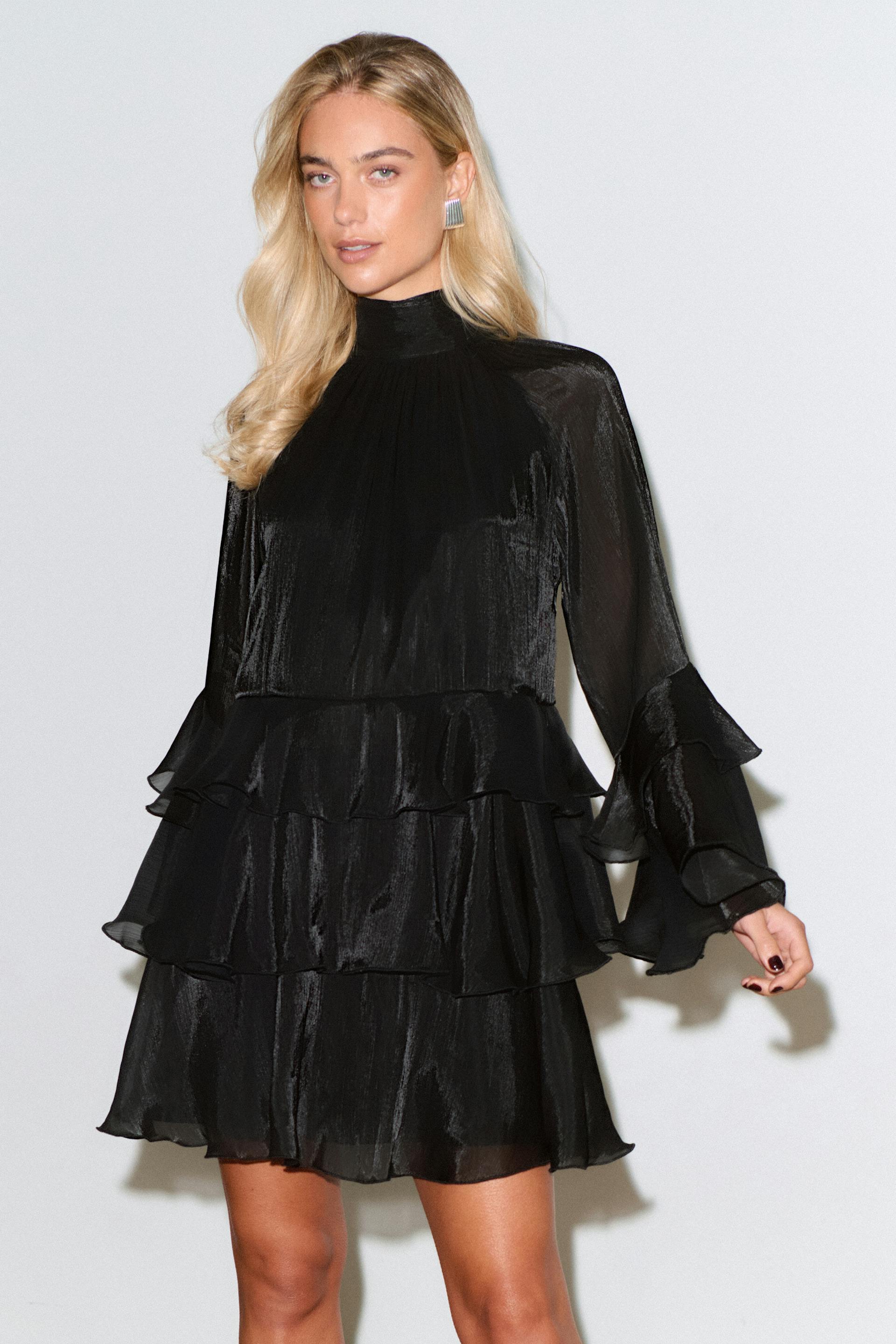 Zwarte glanzende minijurk met hoge hals, lange mouwen met ruches, manchetten met ruches en een rok met drie rijen ruches.