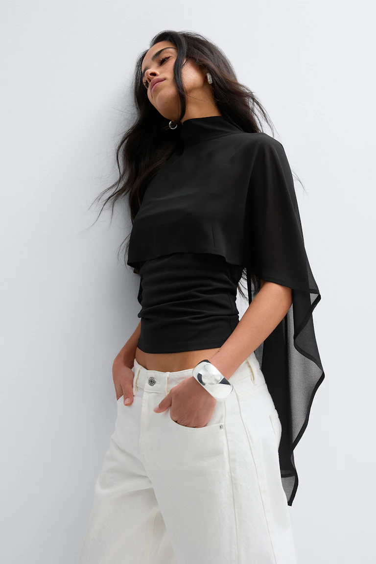 Schwarzes, tailliertes Oberteil mit Mokkakragen und aufgesetztem durchsichtigem Cape, weiße Hose mit weitem Bein und elastischer Taille, silberne Statement-Ohrringe und ein Manschettenarmband.