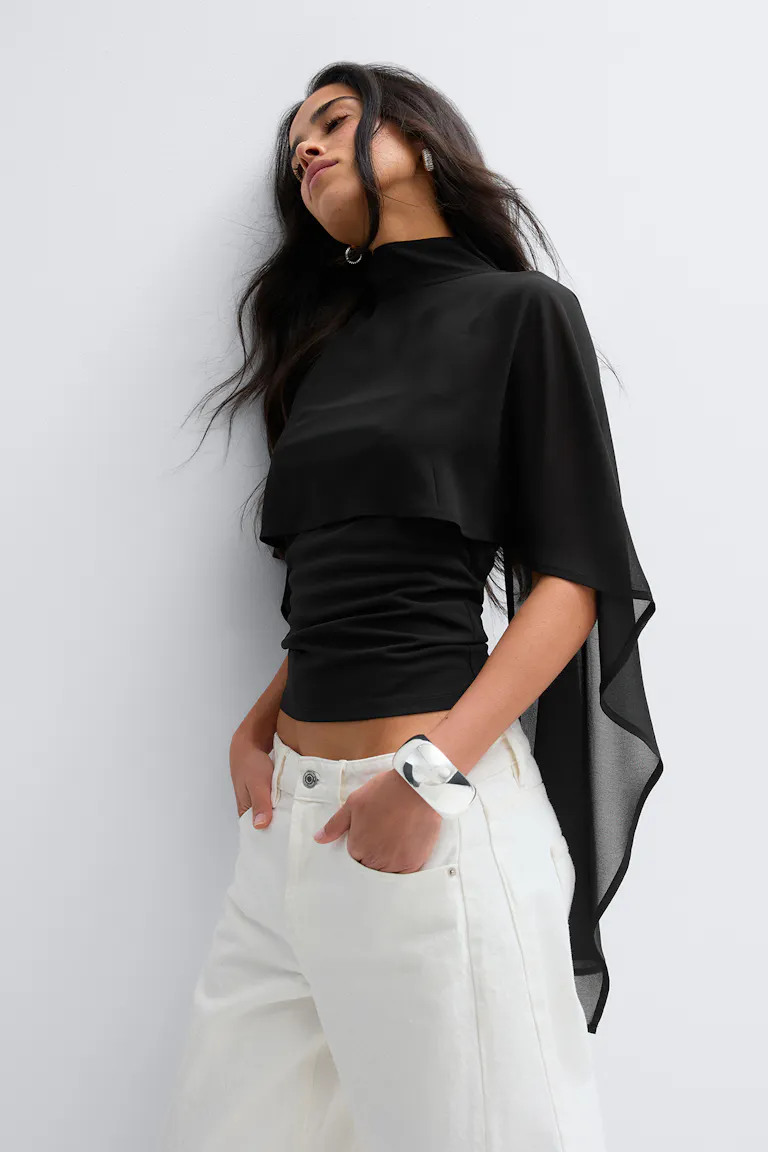 Schwarzes, tailliertes Oberteil mit Mokkakragen und aufgesetztem durchsichtigem Cape, weiße Hose mit weitem Bein und elastischer Taille, silberne Statement-Ohrringe und ein Manschettenarmband.