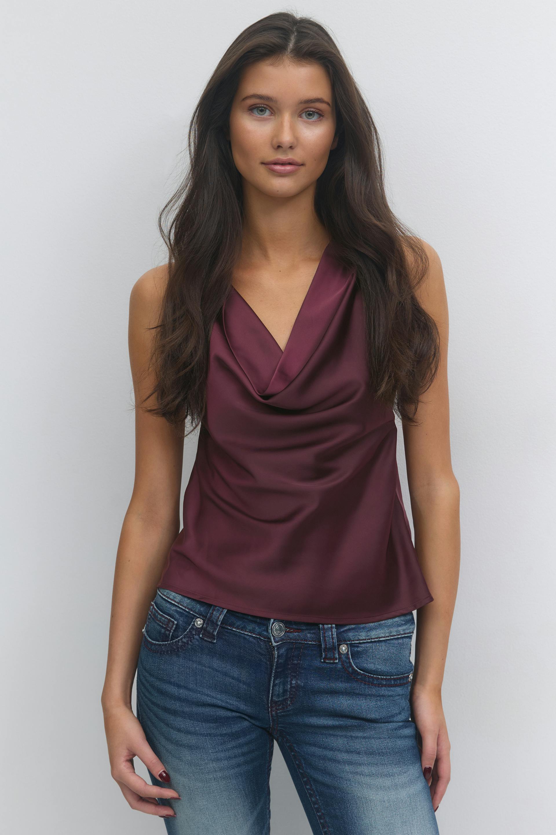 Sleeveless burgundy satin cowl-neck blouse paired with mid-rise faded blue jeans.
