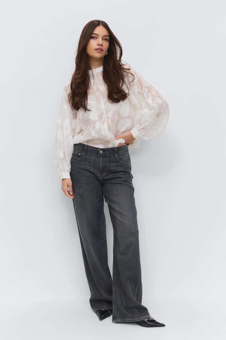 Weiße, durchsichtige, langärmelige Jacquard-Bluse mit hohem Ausschnitt und voluminösen Ärmeln; schwarze, verblasste, hoch taillierte Jeans mit weitem Bein; schwarze, spitze Lederpumps.