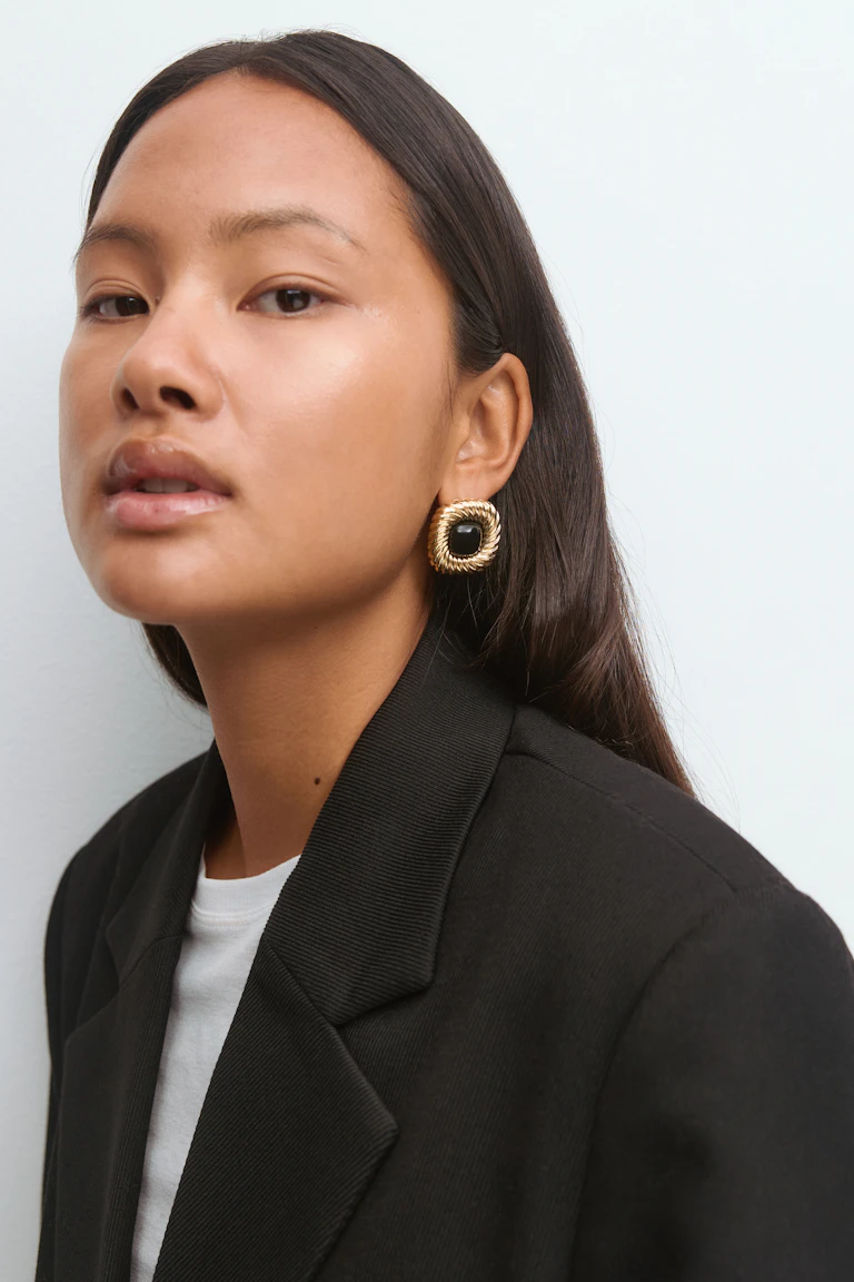 Sort skræddersyet blazer over en hvid T-shirt med rund hals, sammen med firkantede øreringe i guld med rebstruktur og sorte sten i midten.