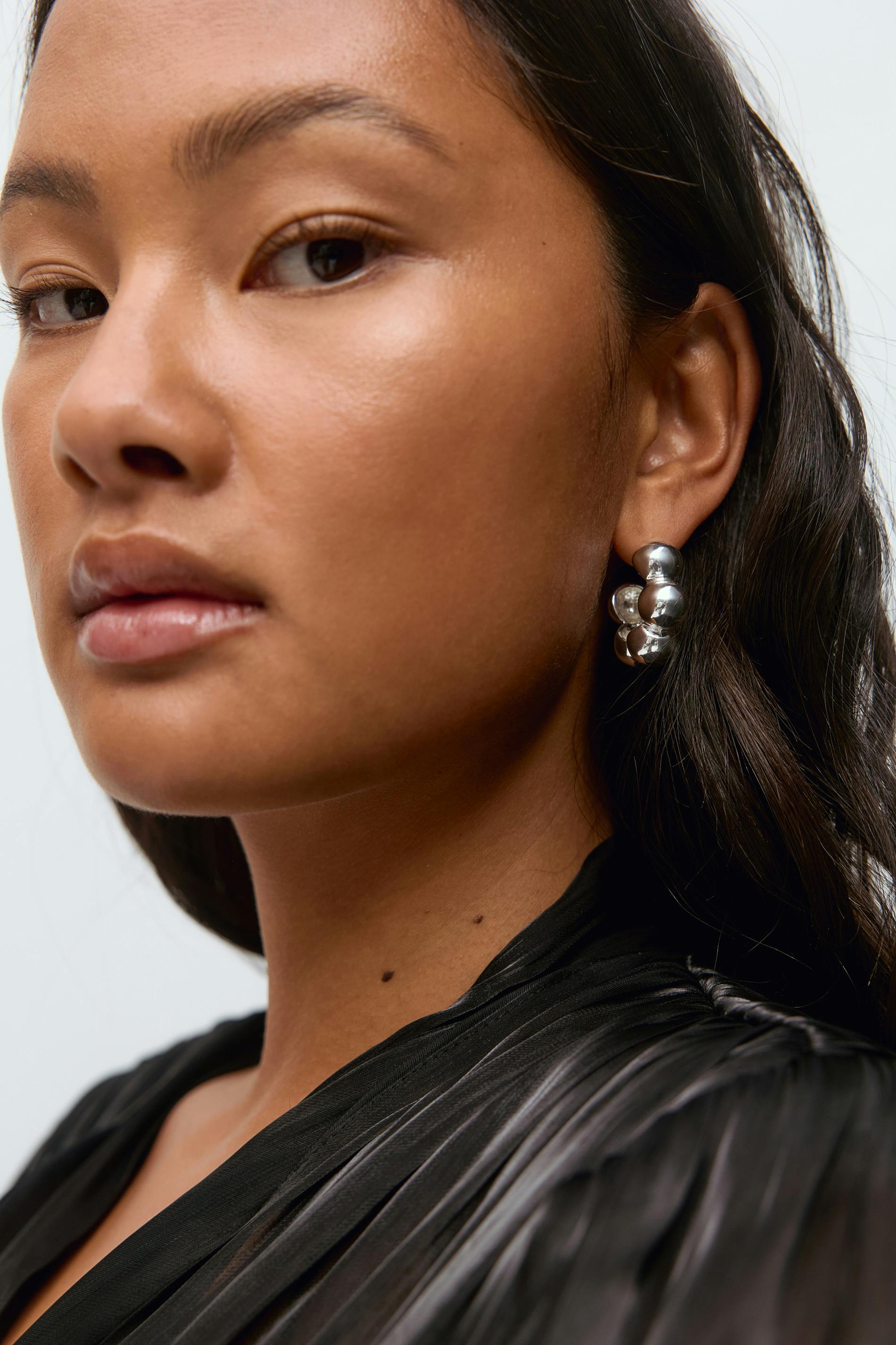 Sie trägt eine schwarze, plissierte Bluse mit dezentem Glanz und silberne Bügelohrringe.